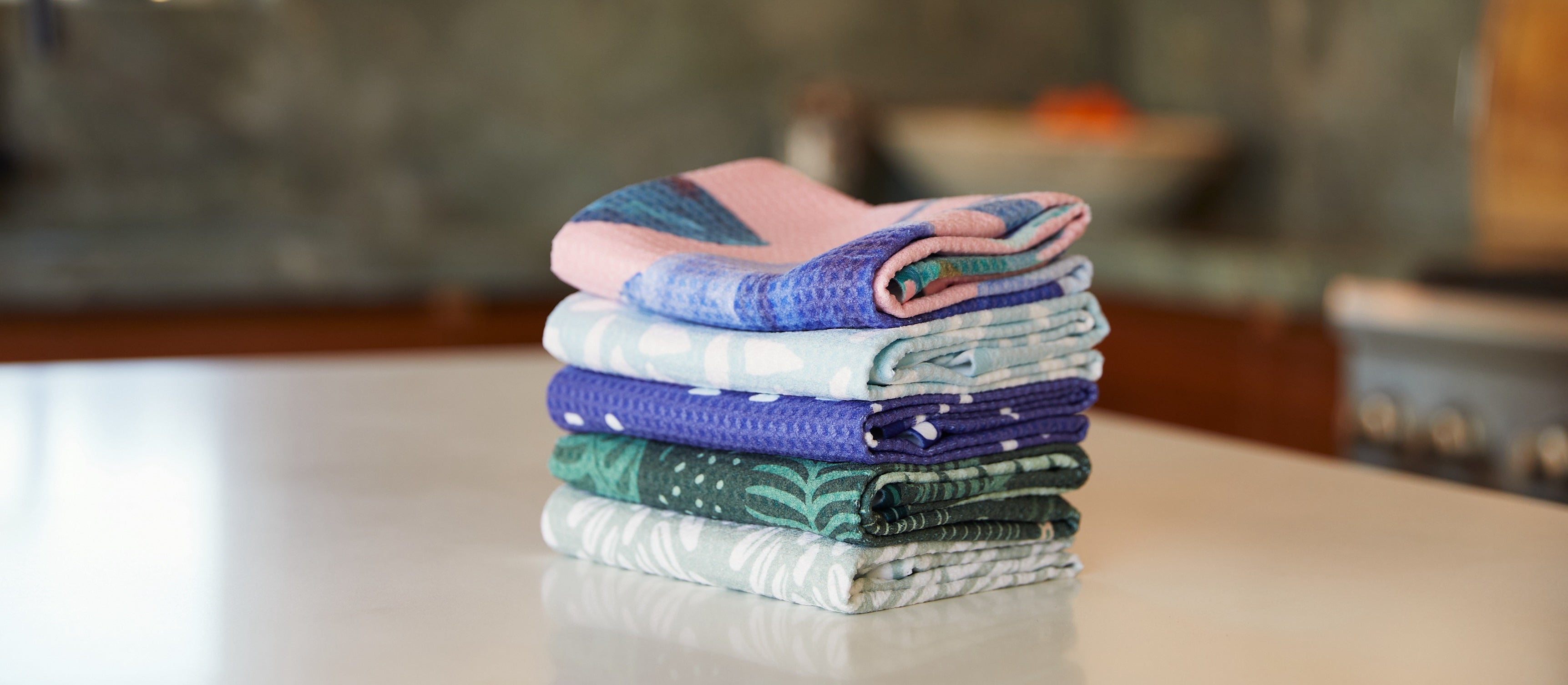 A stack of folded, colorful kitchen towels with various patterns sits on a smooth white countertop in a kitchen with a blurred background.