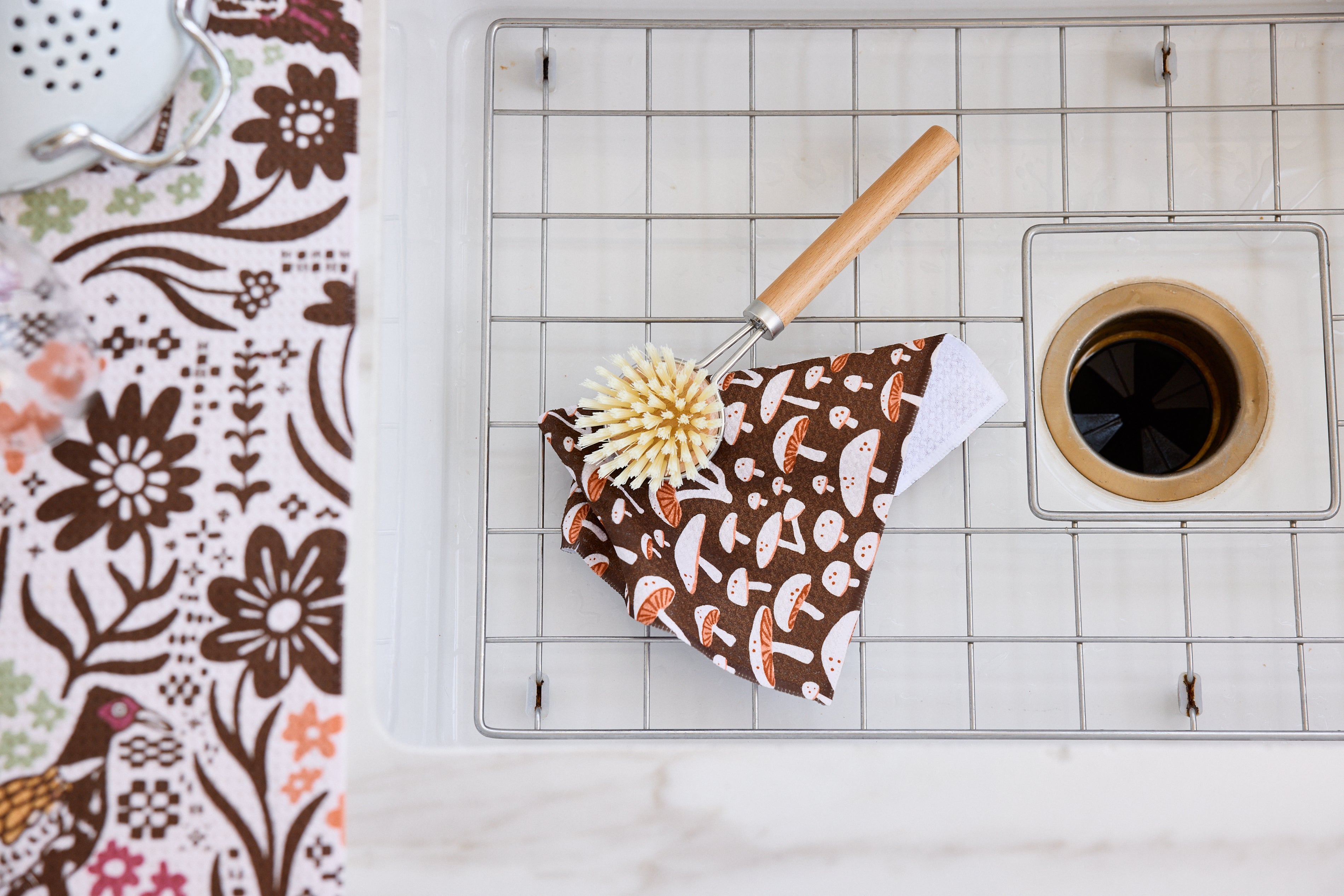A kitchen sink with a brown mushroom-patterned cloth and a wooden-handled scrub brush placed on the metal rack. Part of a floral-patterned cloth is visible on the countertop.