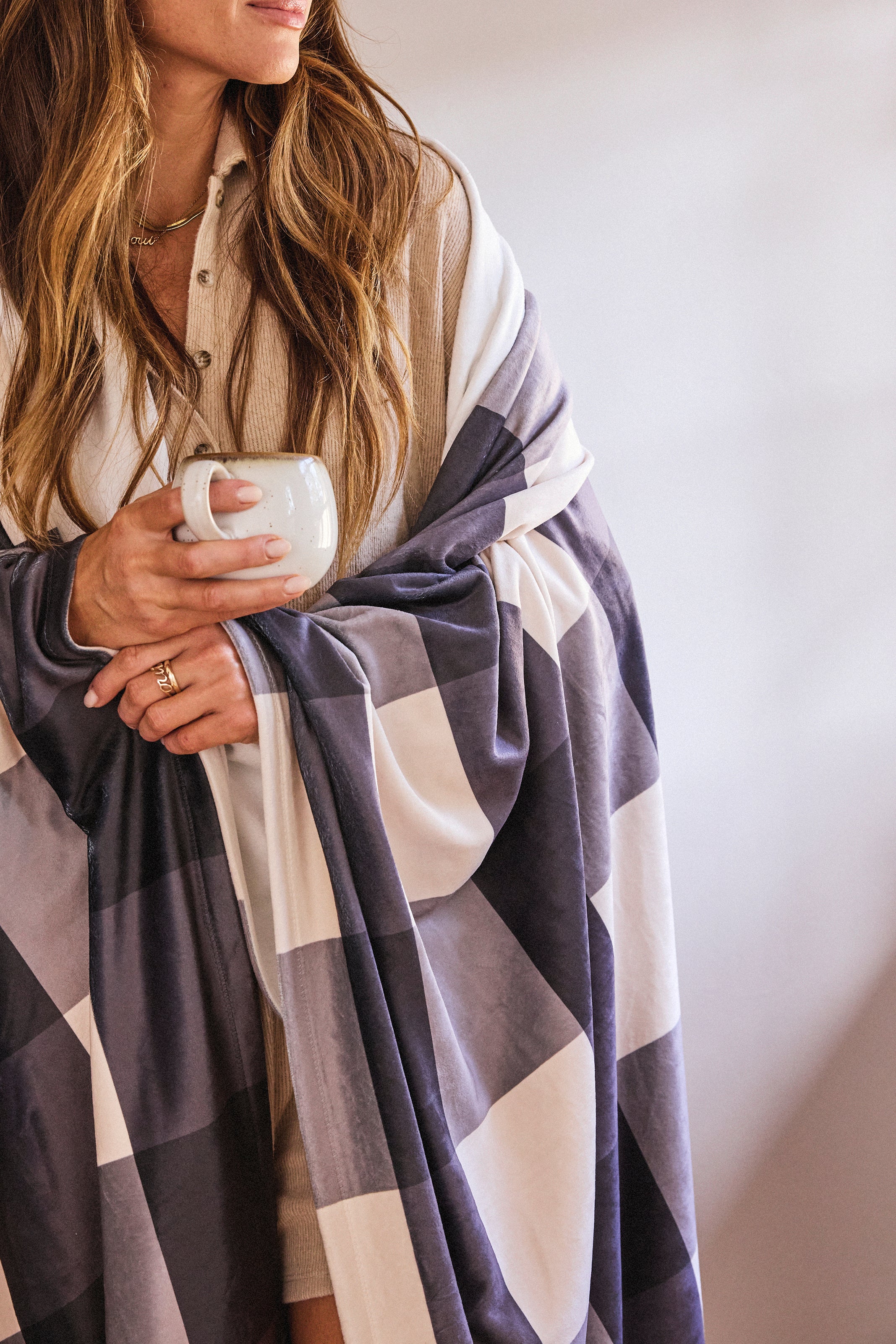 A woman with long hair, wearing a beige button-up, stands against a softly lit background, wrapped in Geometry’s Snowy Gingham blanket made from recycled materials. She holds a white mug and has manicured nails.