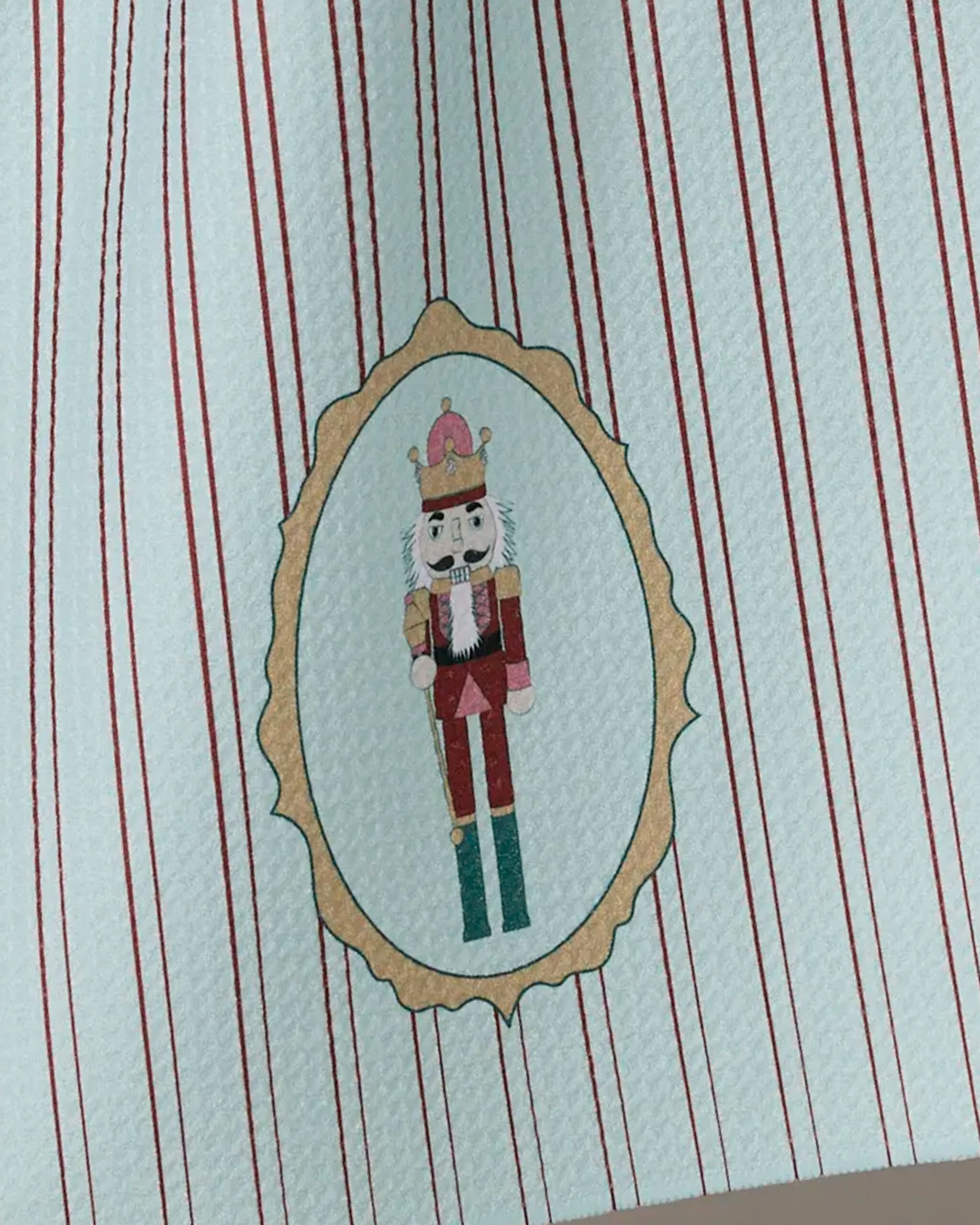 The Geometry "Nutcracker Intermission" tea towel features red and white vertical stripes and a central gold-framed oval with an illustrated nutcracker in a red and green uniform crowned at the center.