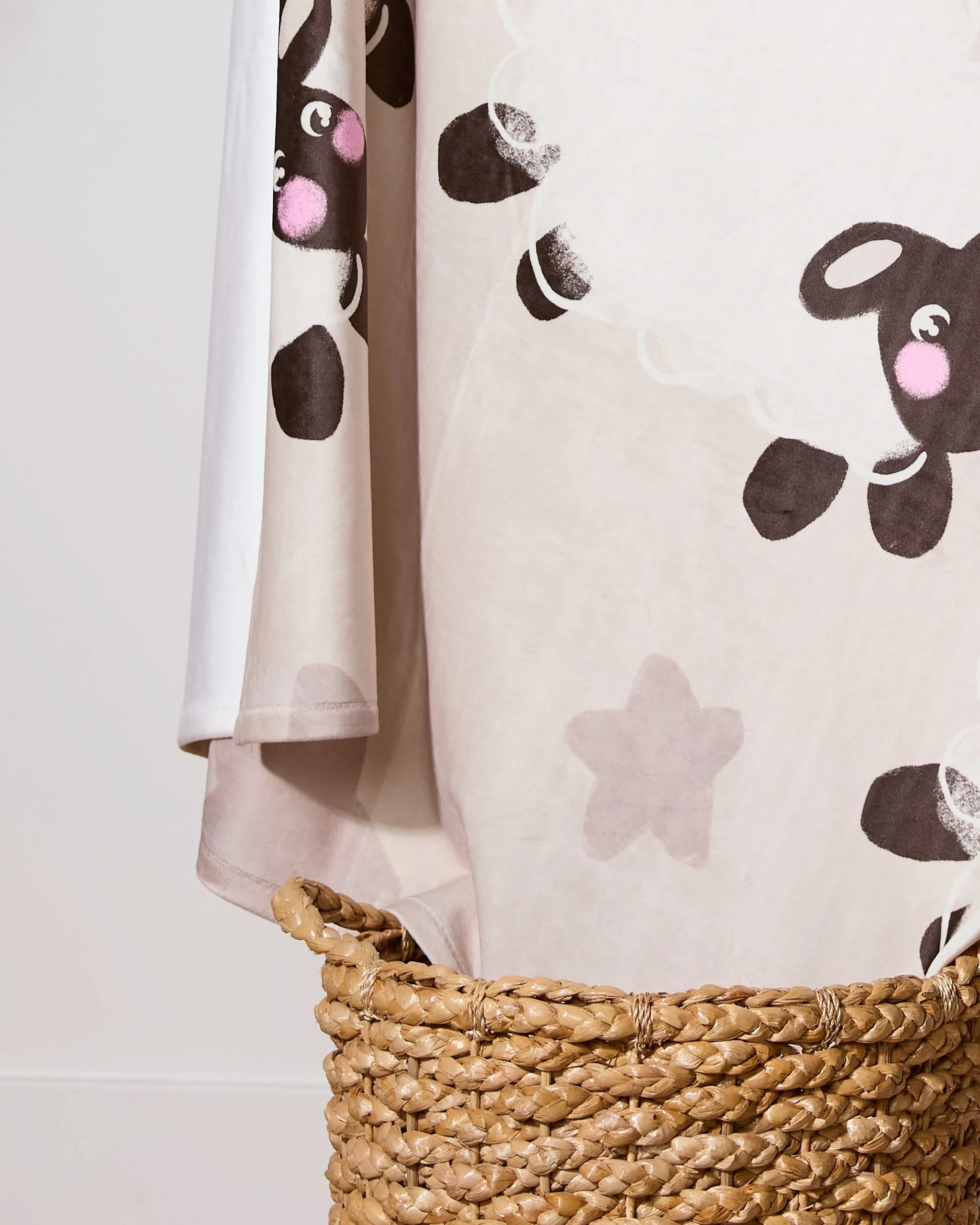 The Geometry Counting Sheep throw blanket, made from cozy recycled materials and adorned with a cute black sheep cartoon design and pale stars, drapes over the side of a woven basket against a light background.