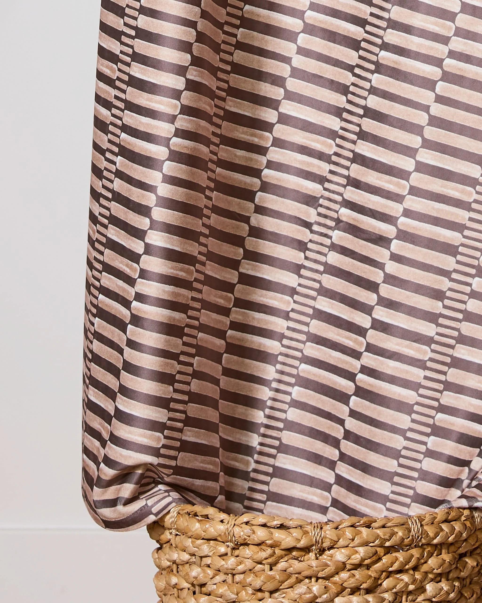 A close-up of Geometrys Light Ridge recycled blanket, featuring a brown and beige geometric pattern, draped over a woven natural-colored basket against a neutral background.