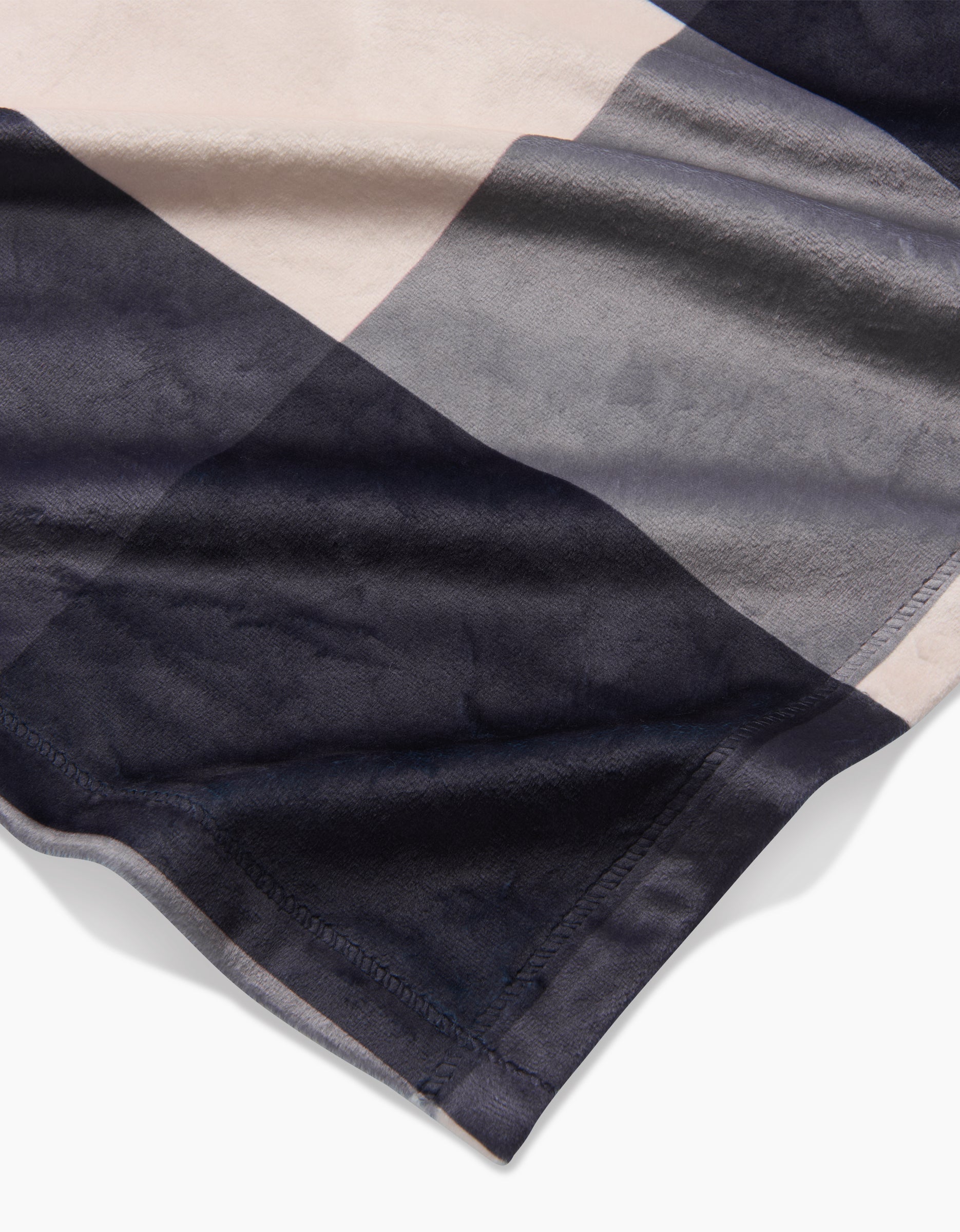 A close-up of Geometry’s Snowy Gingham blanket, made from recycled materials, features large overlapping rectangular blocks in beige, gray, and dark blue, showcasing its silky softness and detailed edge stitching.