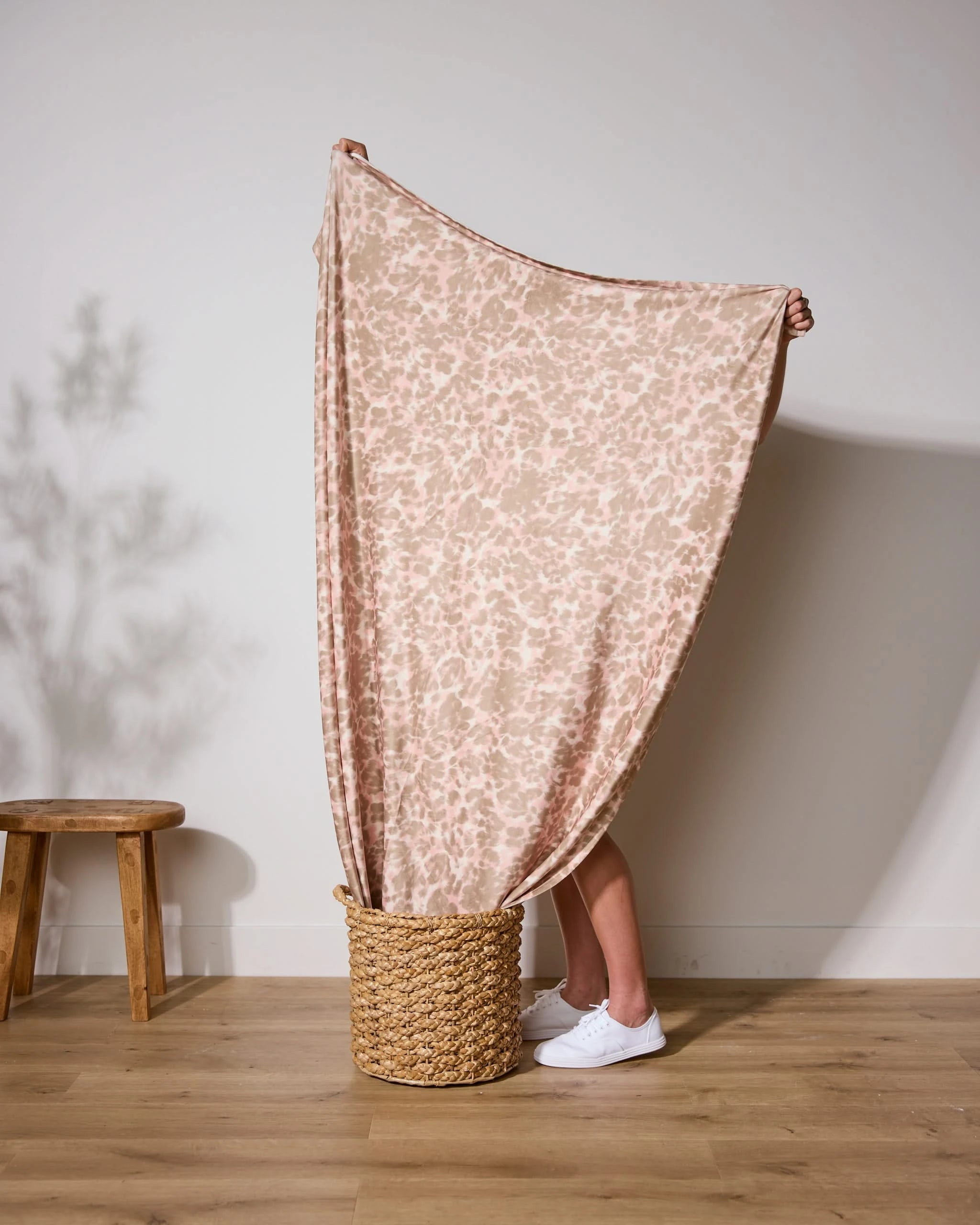 Standing in a woven basket, a person holds up the Soft Umber blanket by Geometry, its light pink fabric covering their upper body and face. Only their legs and white sneakers are visible. A wooden stool sits nearby against a plain wall.