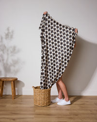 A person stands barefoot on a wooden floor, mostly hidden behind Geometry’s Highland Lullaby blanket in a large black-and-white houndstooth pattern. Only their lower legs and feet appear near a woven basket and wooden stool.