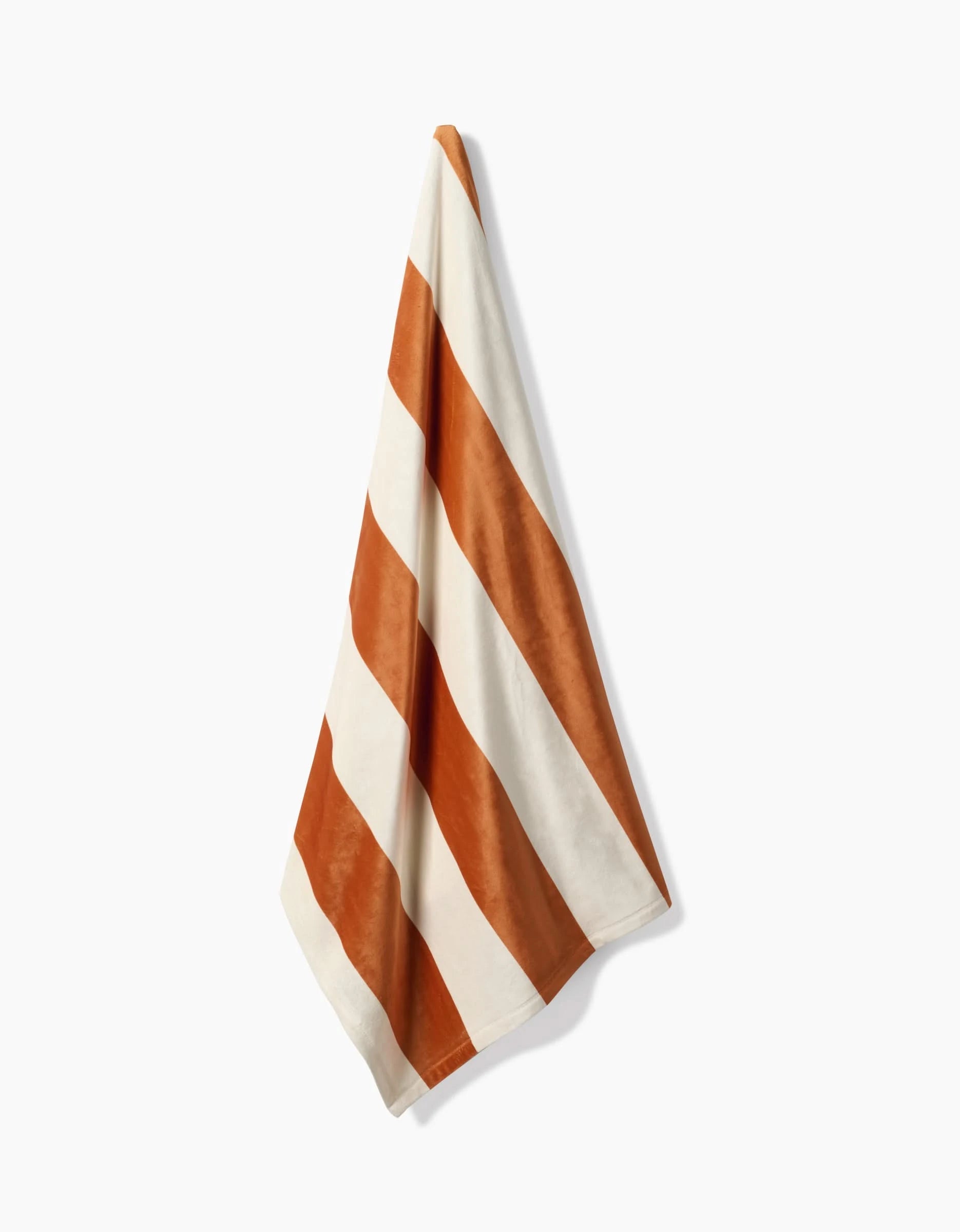 The Geometry Radiance towel, featuring bold diagonal orange and white stripes and made from recycled materials, hangs against a plain white background.