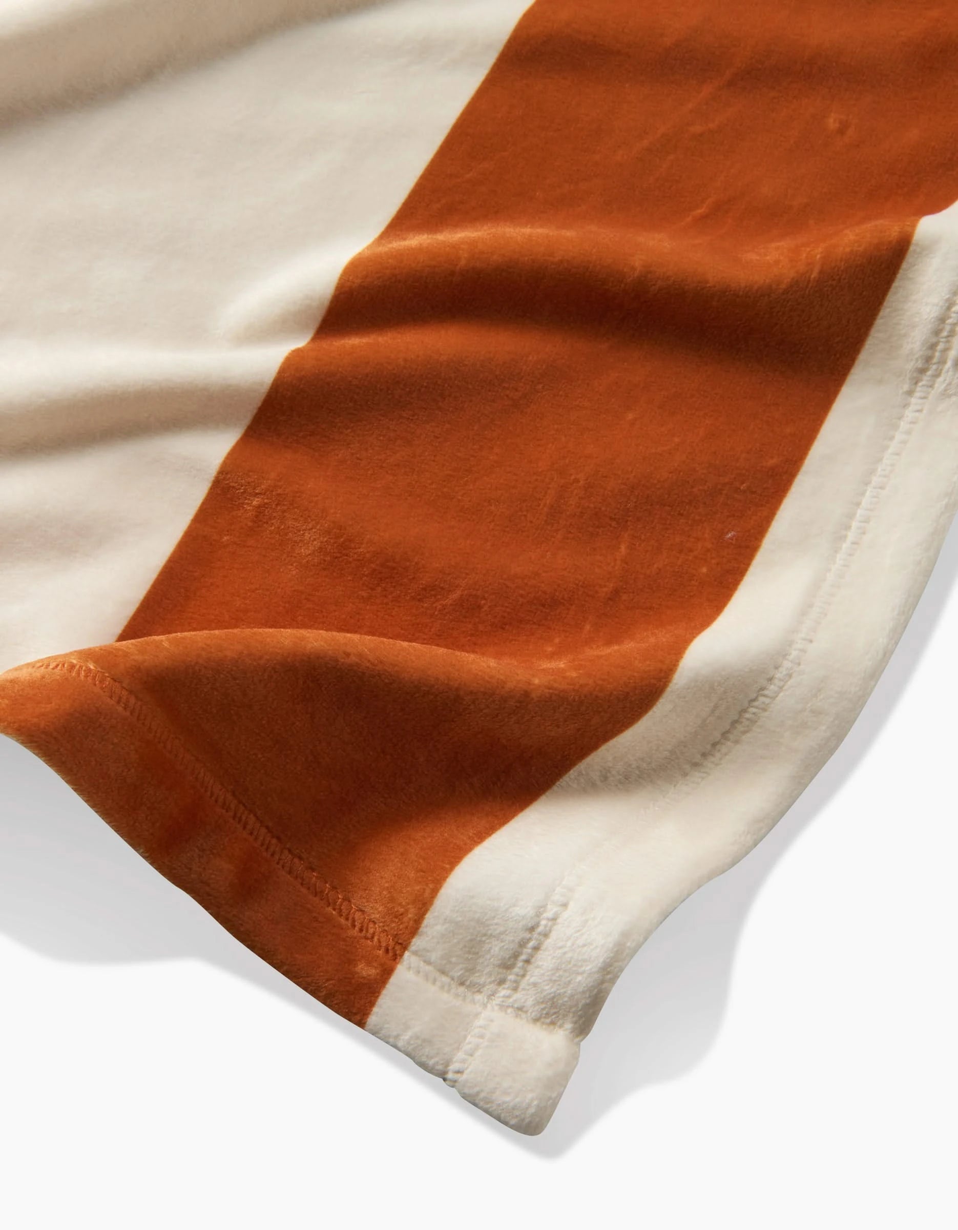 A close-up of Geometry’s Radiance Butter Blanket, crafted from recycled materials, featuring a wide rust-orange stripe and an off-white stripe, with visible stitching along the edge and a slight fold in the silky-soft fabric.