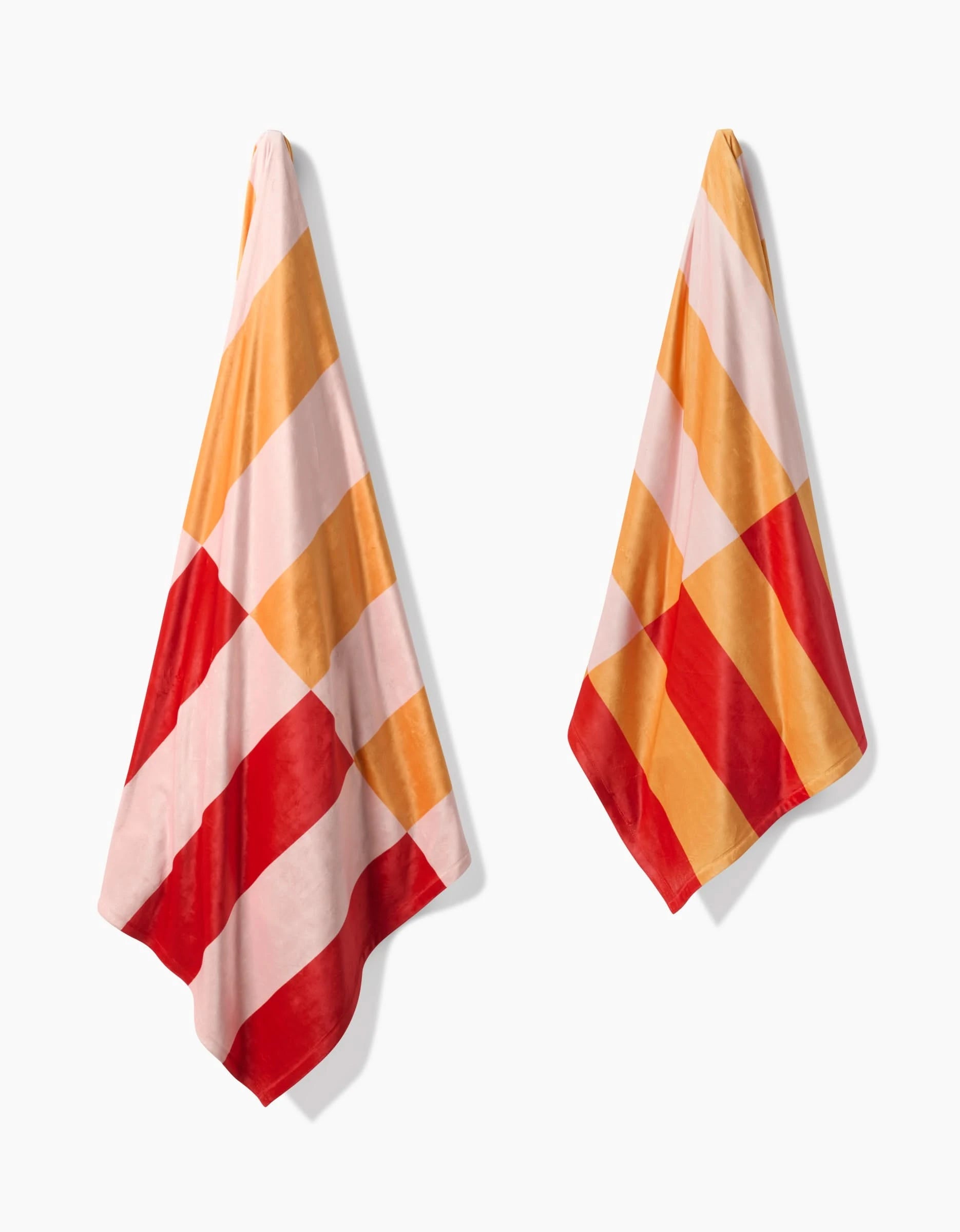 Two Geometry Geometric Sunset towels with wide diagonal stripes in pink, orange, and red hang on hooks against a white background, crafted from recycled materials for extra silky softness.