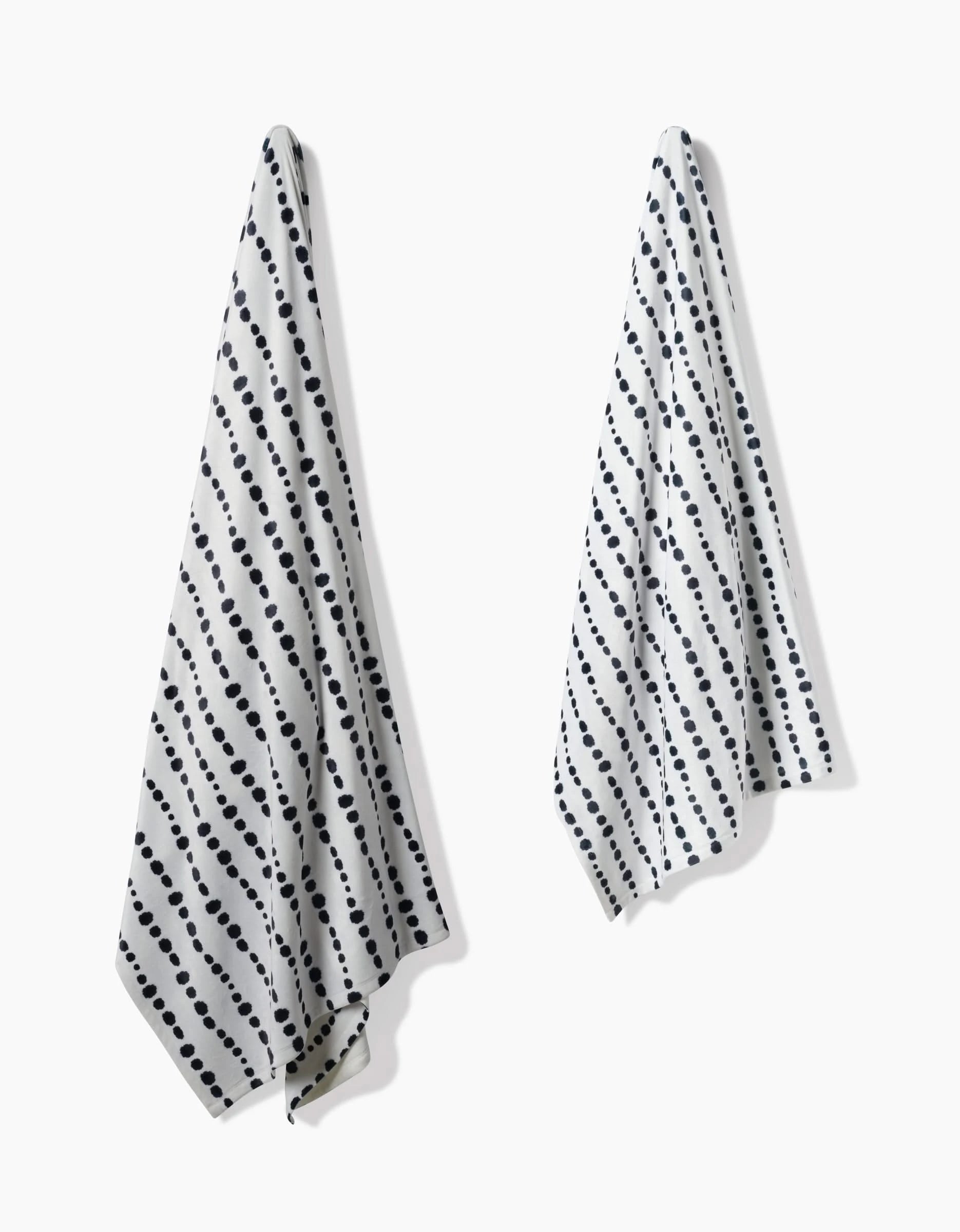 Two Geometry Ink Dots white towels with diagonal black polka dot stripes hang separately on a white wall, casting soft shadows—made with the same recycled material care as our eco-friendly blankets.