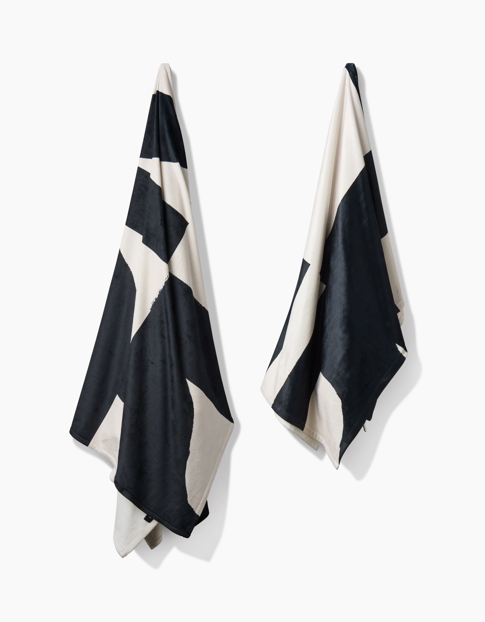Two Lined Square towels by Geometry, featuring bold black and white geometric patterns, hang against a plain white backdrop. Made from post-consumer recycled materials, they showcase contrasting triangles and a silky soft texture.