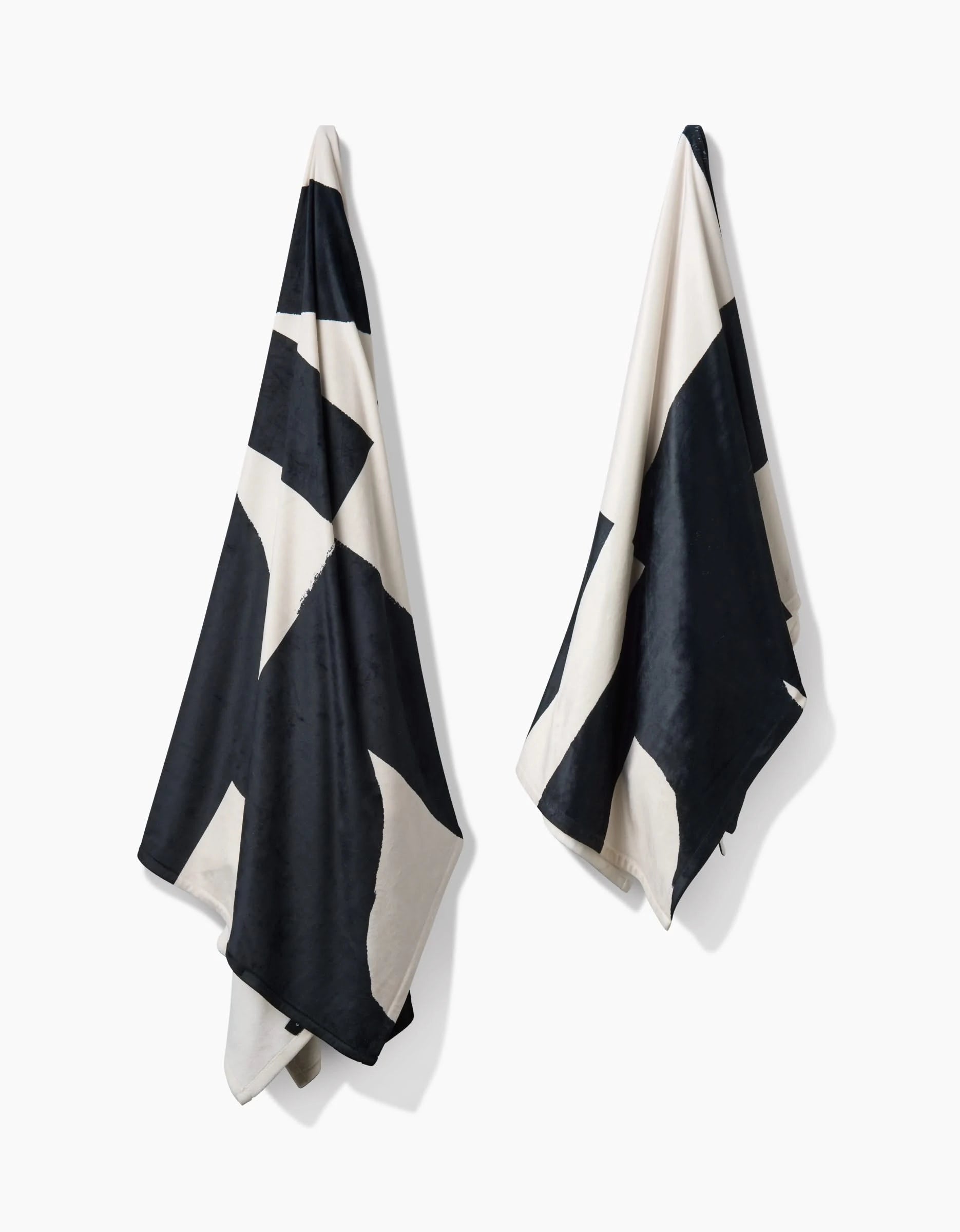 Two Lined Square towels by Geometry, featuring bold black and white geometric patterns, hang against a plain white backdrop. Made from post-consumer recycled materials, they showcase contrasting triangles and a silky soft texture.