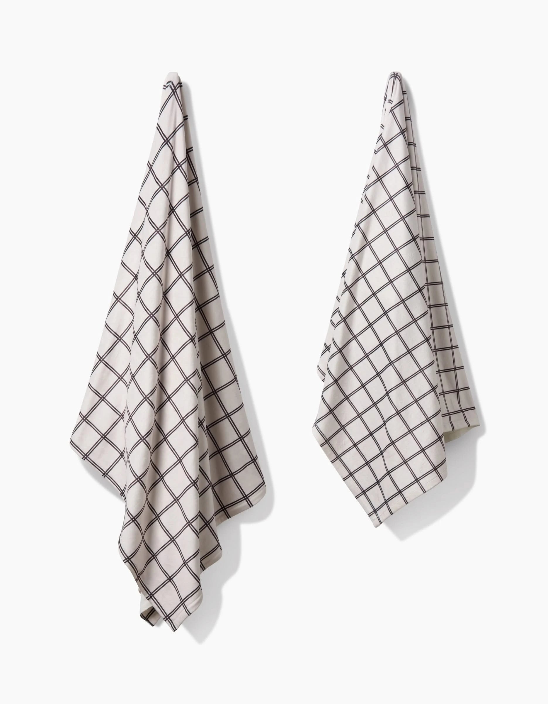 Two Moon kitchen towels by Geometry, featuring a beige color with a black grid pattern and made from recycled materials, hang side by side on hooks against a white background, casting soft shadows.