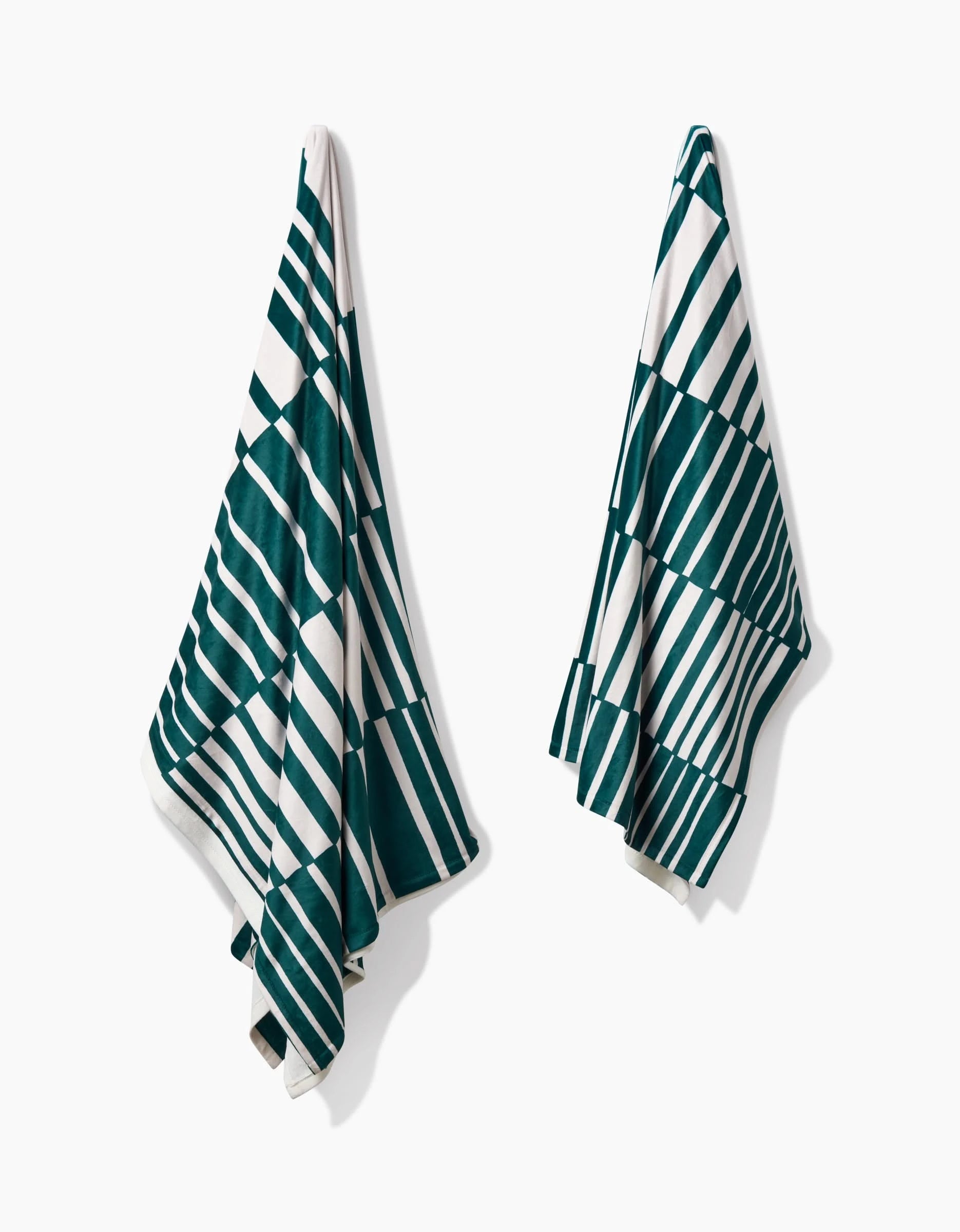 Two Verdelia towels by Geometry, featuring green and white alternating horizontal and diagonal stripes, hang separately on a white wall. Each towel is crafted from recycled materials.