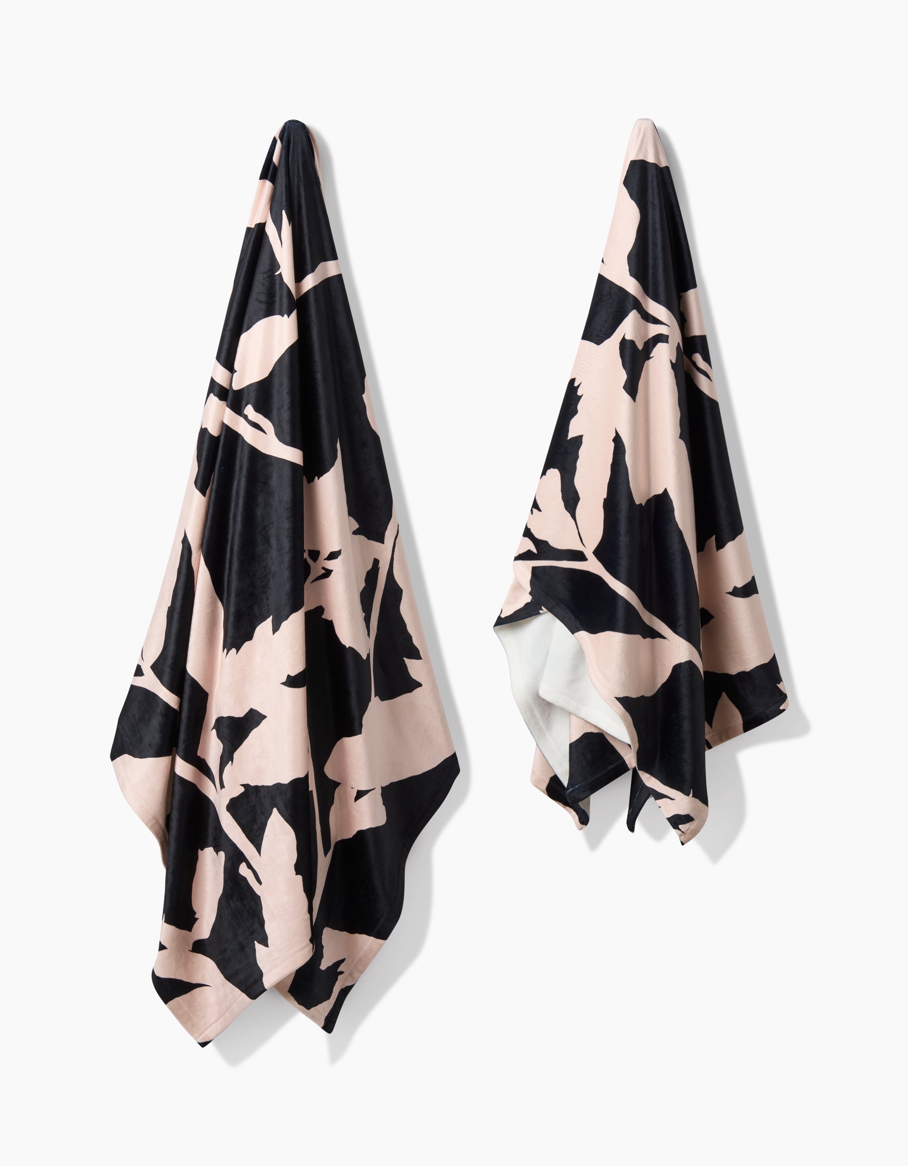 Two Geometry Pam towels with modern abstract black and light pink patterns hang on hooks against a white background. Made from recycled materials, they feature soft, draped fabric and asymmetric botanical-style designs.