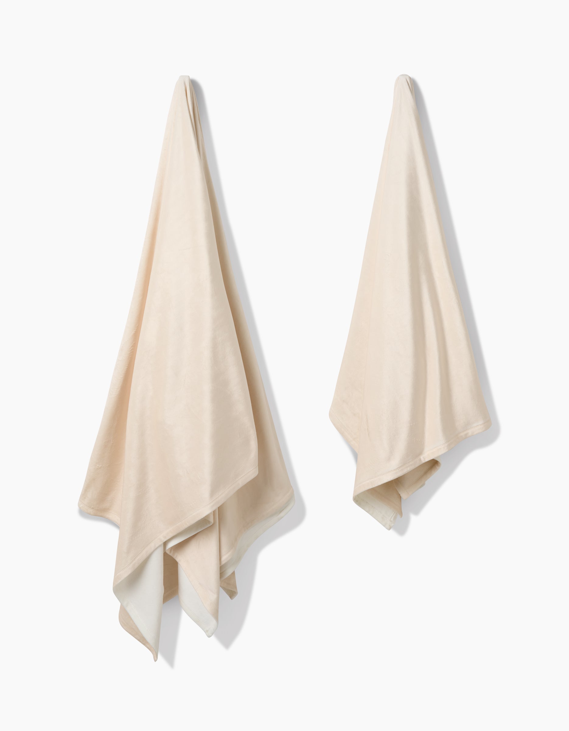 Two beige Soft Glow towels by GEOMETRY, made from eco-friendly materials, hang separately on hooks against a plain white background. The towels look soft and plush with natural folds and slightly uneven edges.