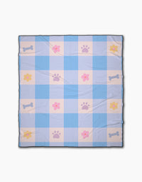 The Blue Sky Picnic Dreams blanket by Letters By Shells is a light blue and white checkered beach blanket with pink paw prints, yellow and pink flowers, and blue bones in a repeating pattern.