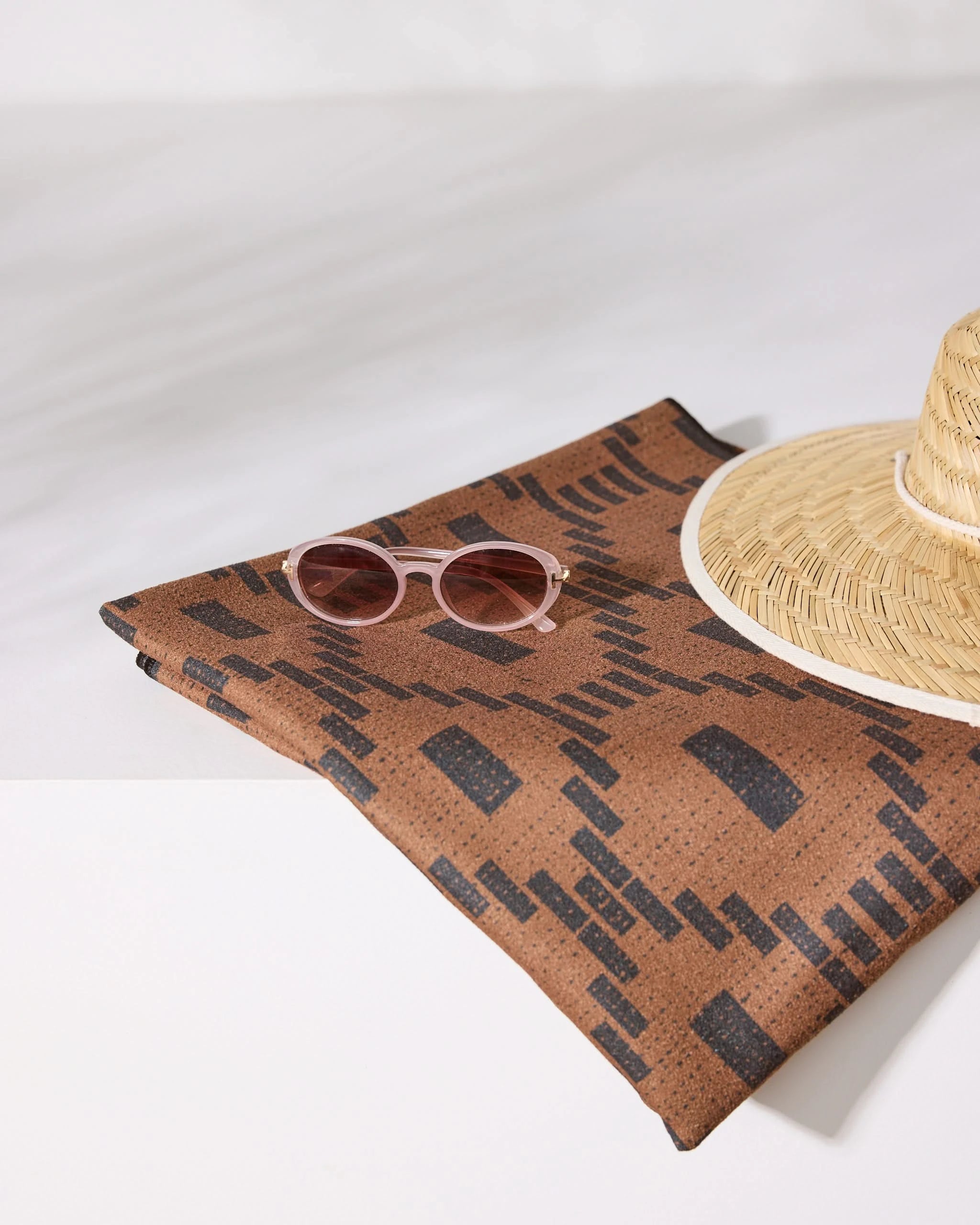 A straw hat and pink sunglasses rest on a folded Geometry Caramel Kiln Micro-loop Beach Blanket, showcasing its brown and black pattern against a crisp white background.