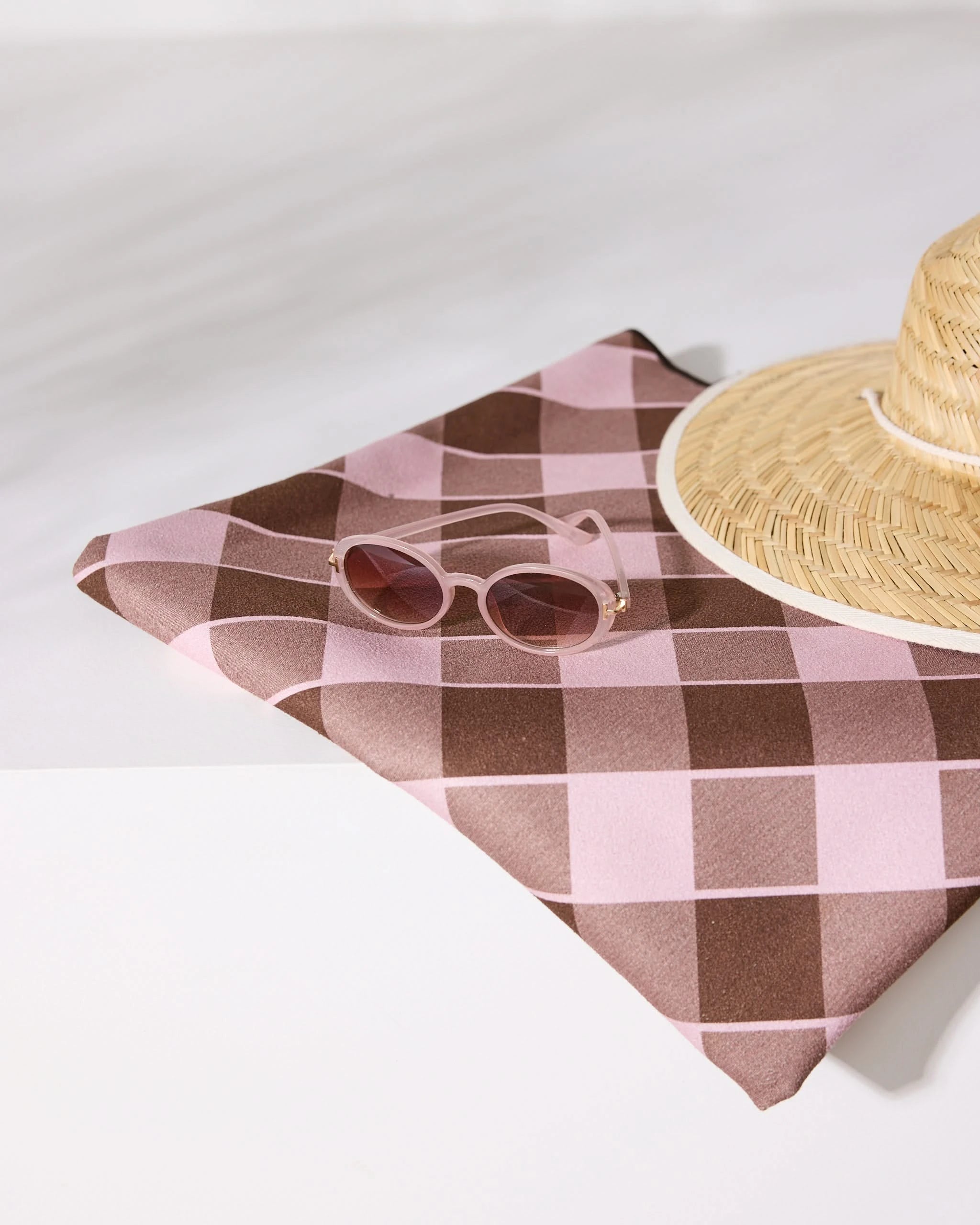 A pair of pink sunglasses and a straw hat sit atop the Geometry Choco Berry pink and brown checkered quick-drying beach blanket, neatly folded on a white surface with soft shadows.