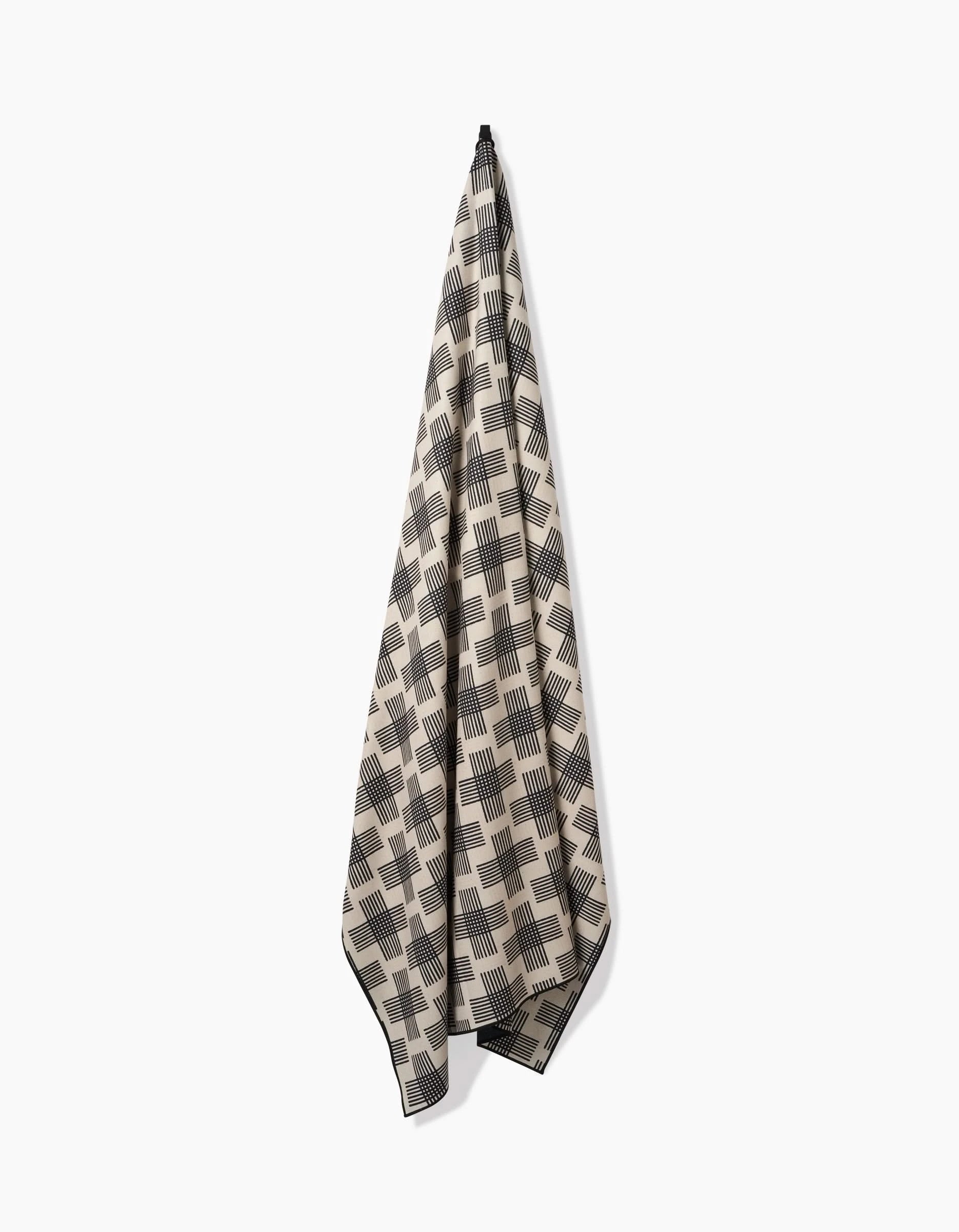 The Rosa Kichline Cross Hatch towel, made from recycled materials, hangs from a single hook against a white background. It features a beige and black geometric square and stripe design for an eco-friendly, stylish touch.
