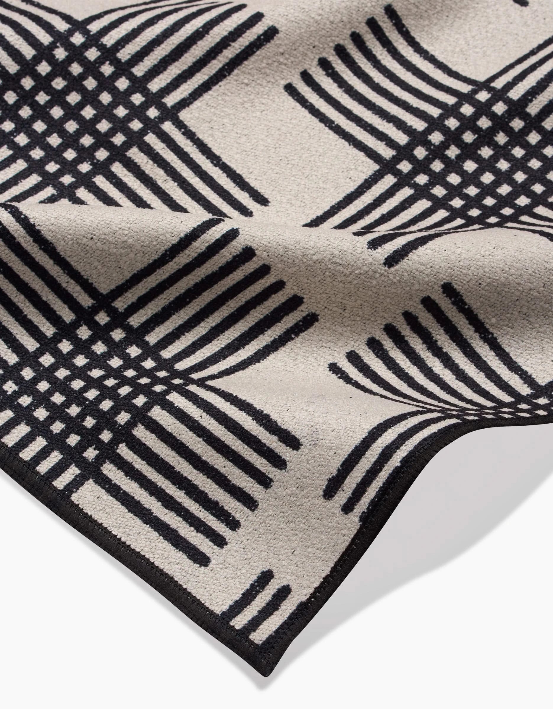 The Cross Hatch Beach Blanket by Rosa Kichline features a beige recycled fabric with a black geometric intersecting line design forming curved squares, shown slightly folded to highlight its textured surface and edge detail.