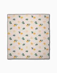 The Sara Mari Freshly Picked beach blanket is cream-colored, quick-drying, and made from recycled materials. It features a repeating pattern of yellow lemons, lemon slices, and green leaves with slight visible wrinkles when spread out.