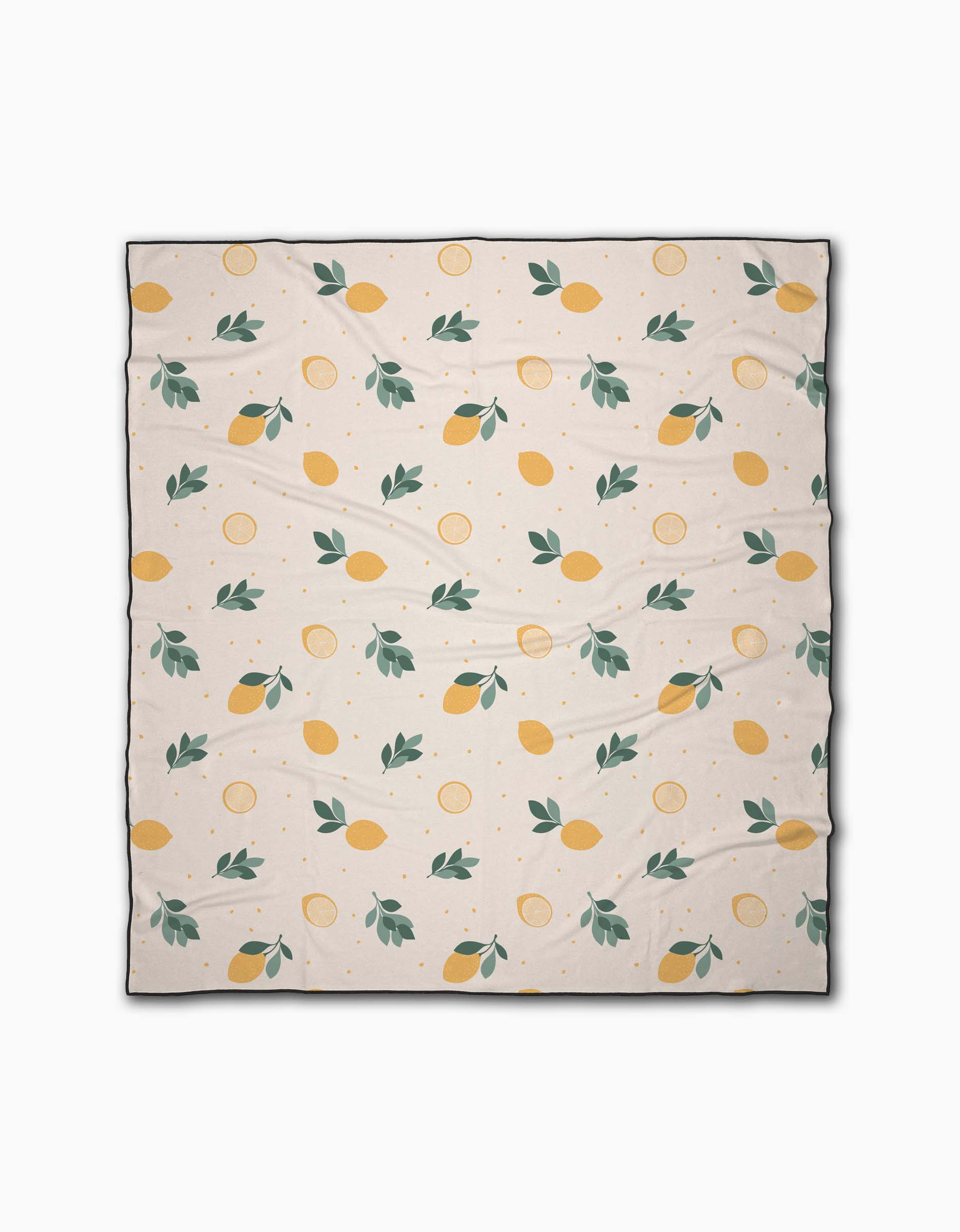 The Sara Mari Freshly Picked beach blanket is cream-colored, quick-drying, and made from recycled materials. It features a repeating pattern of yellow lemons, lemon slices, and green leaves with slight visible wrinkles when spread out.