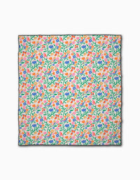 The Primrose Path beach blanket by Charlie Rowan Designs features a colorful, dense floral pattern with green stems and pink, orange, purple, and blue flowers on white. Made from recycled materials with black-edged detailing.