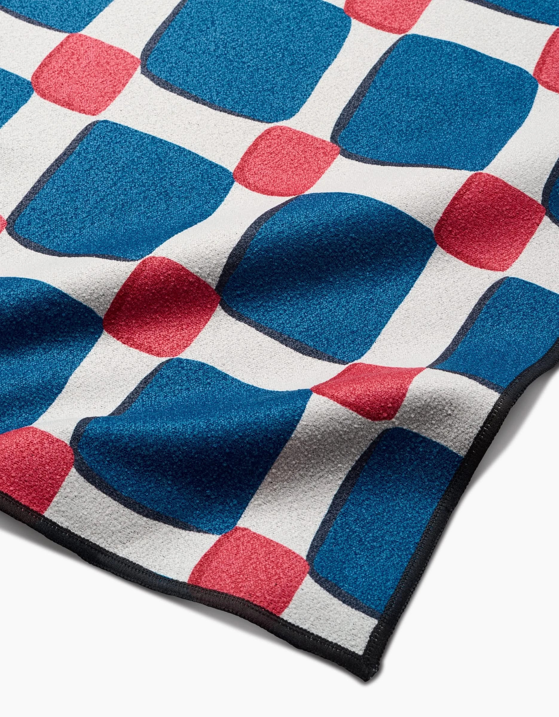 The Box Check Picnic by Hufton Studio is a micro-loop beach blanket with large blue and small red squares on white, black edge trim, and made from quick-drying recycled materials. The fabric is soft and subtly textured.