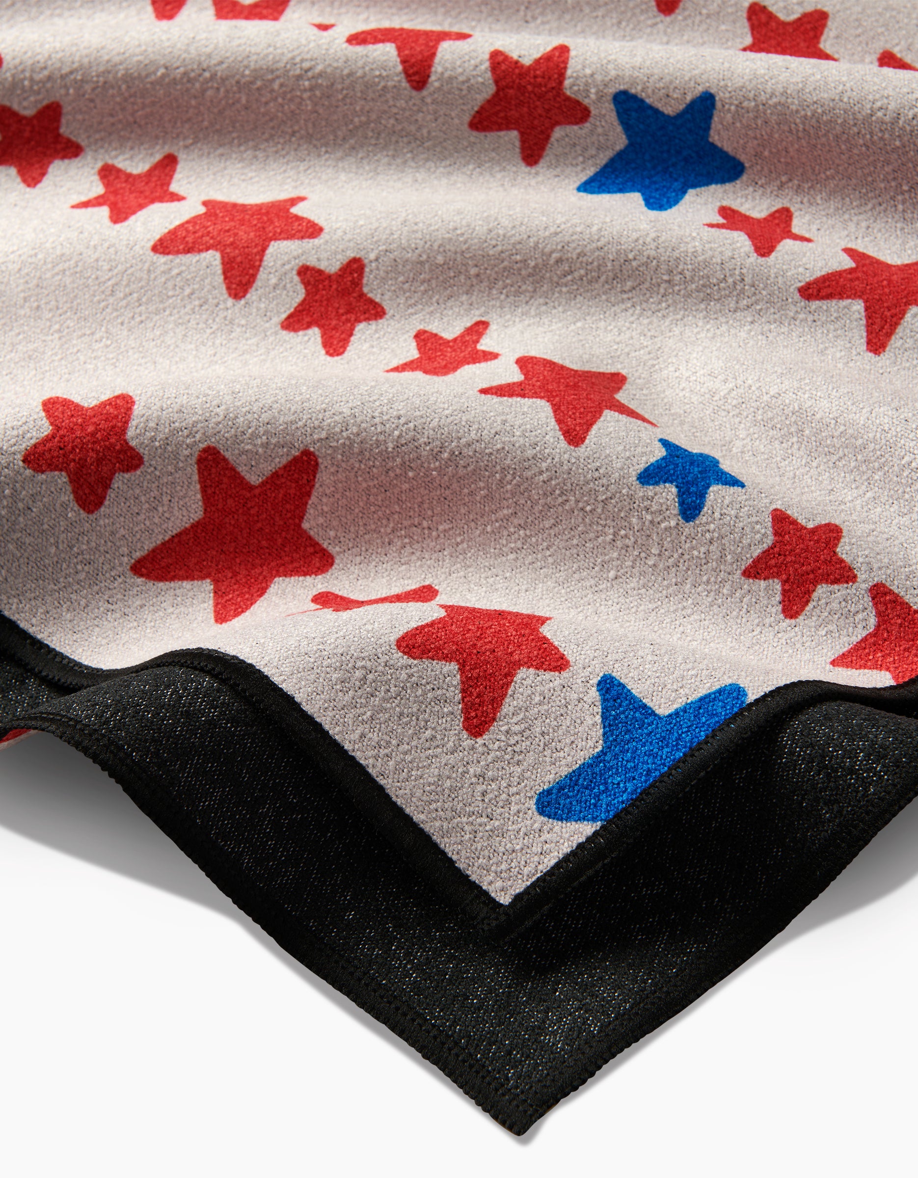 A close-up of Kayla Ann’s Starry Stripes micro-loop beach towel, showcasing red and blue stars on a light background with a black border. Made from quick-drying recycled materials, the fabric features a textured, slightly wavy look.
