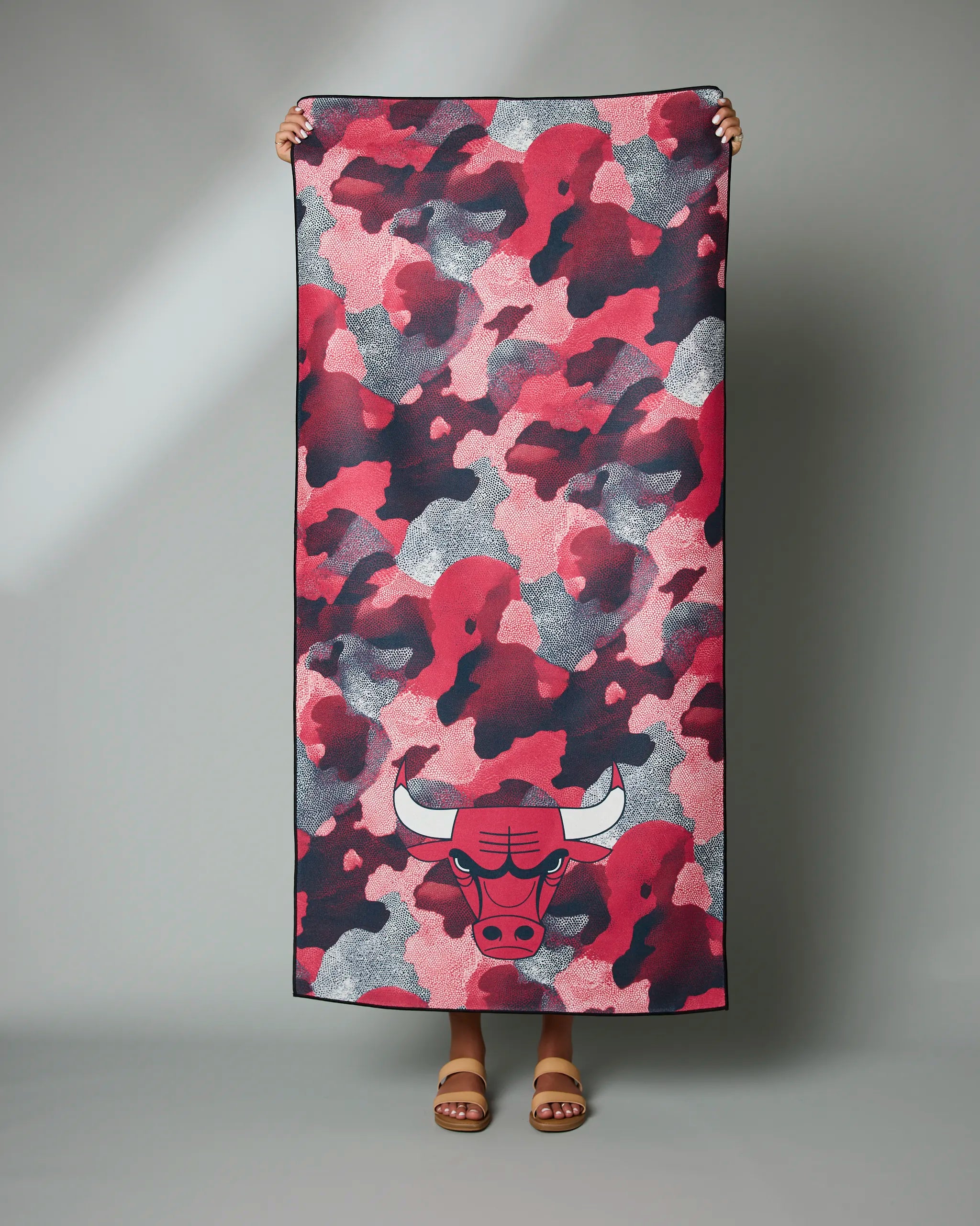 A person holds up the Geometry Bulls Camo towel, featuring a rectangular shape, a red, pink, black, and gray camouflage pattern, and a bull logo near the bottom. Only their hands and feet are visible.