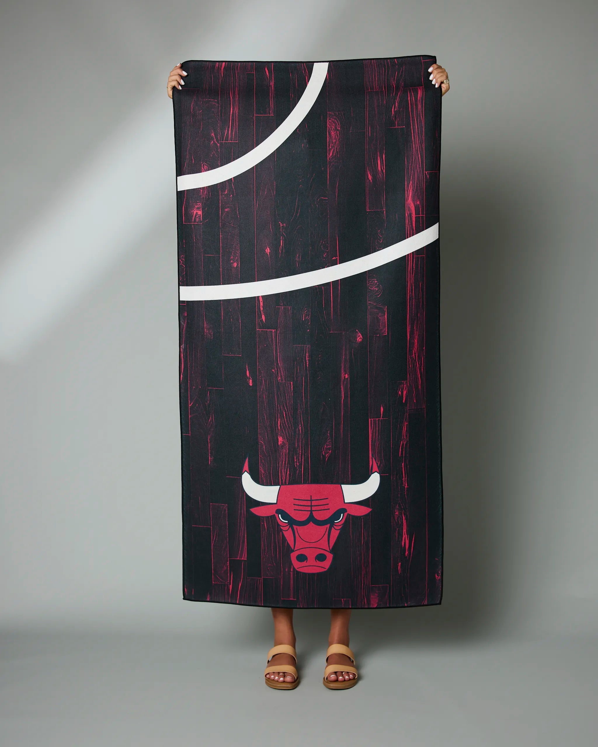 A person displays Geometry’s Bulls Zone rectangular towel featuring the Chicago Bulls logo and a basketball court design on a black-and-red wood-patterned background; only their hands and feet are shown.