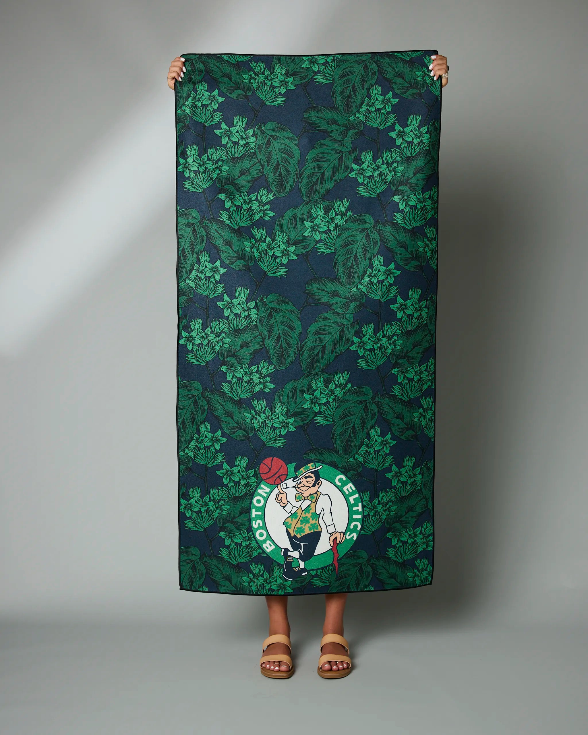 A person holds up the Geometry Celtics Floral towel, featuring green leafy patterns and the Boston Celtics logo in one corner; only their hands and feet are visible.
