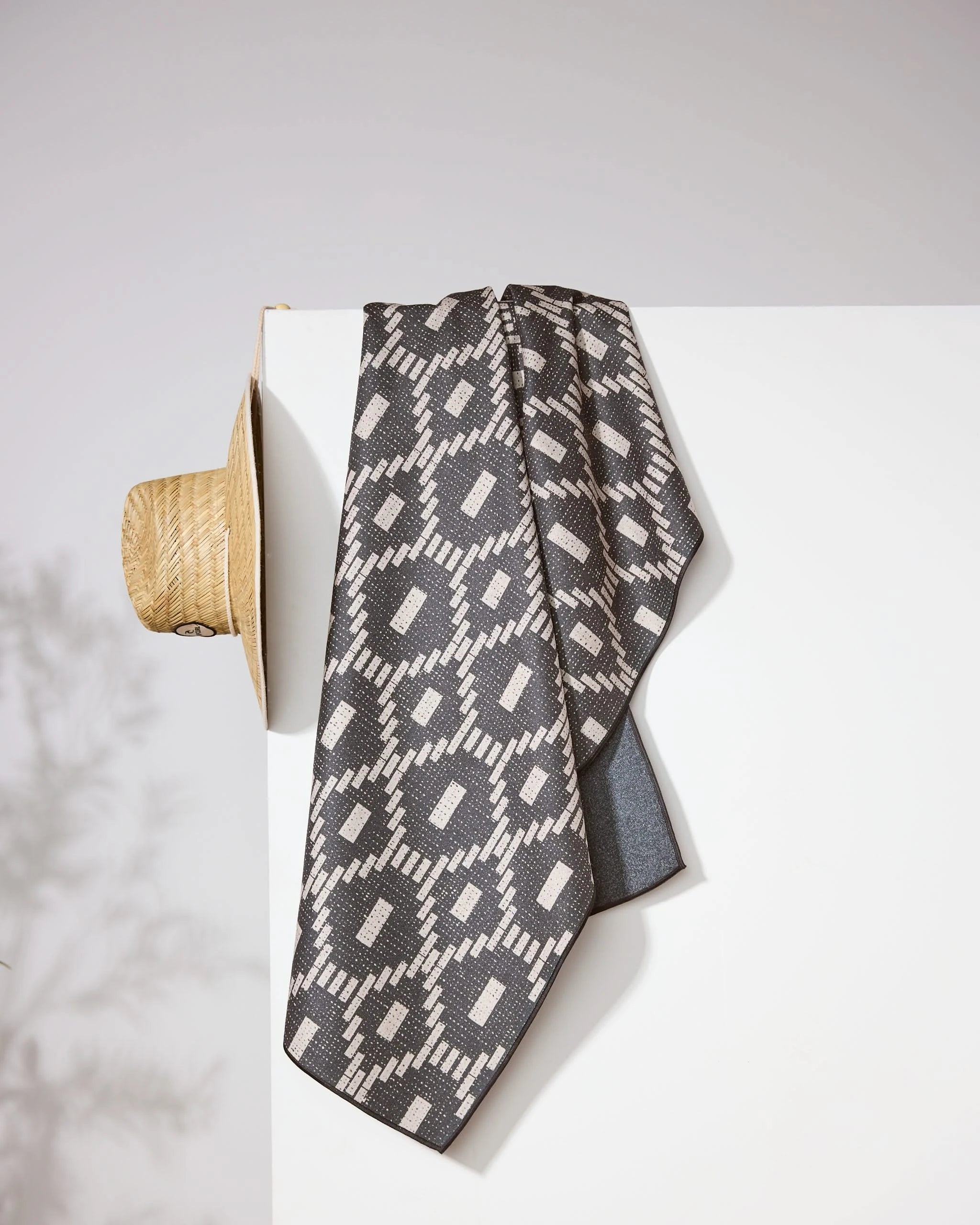 A black and gray Geometry Charcoal Kiln beach towel with geometric patterns drapes over a white wall. A straw hat hangs on the corner, with a subtle plant shadow in the background.