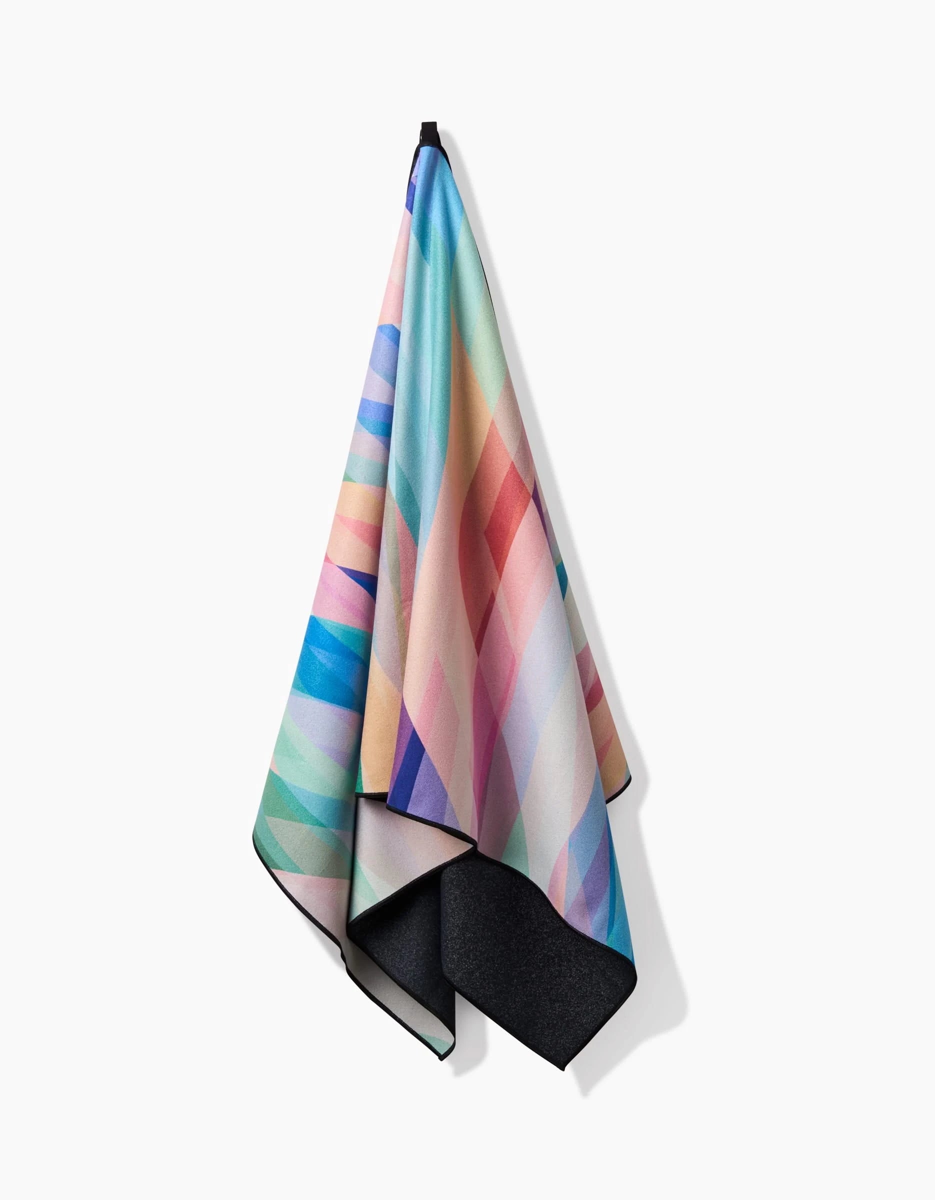 The Rachel Parker Eventide Micro-loop Beach Towel, featuring a pastel geometric pattern in blue, pink, green, and purple with a dark gray underside, hangs on a white wall.