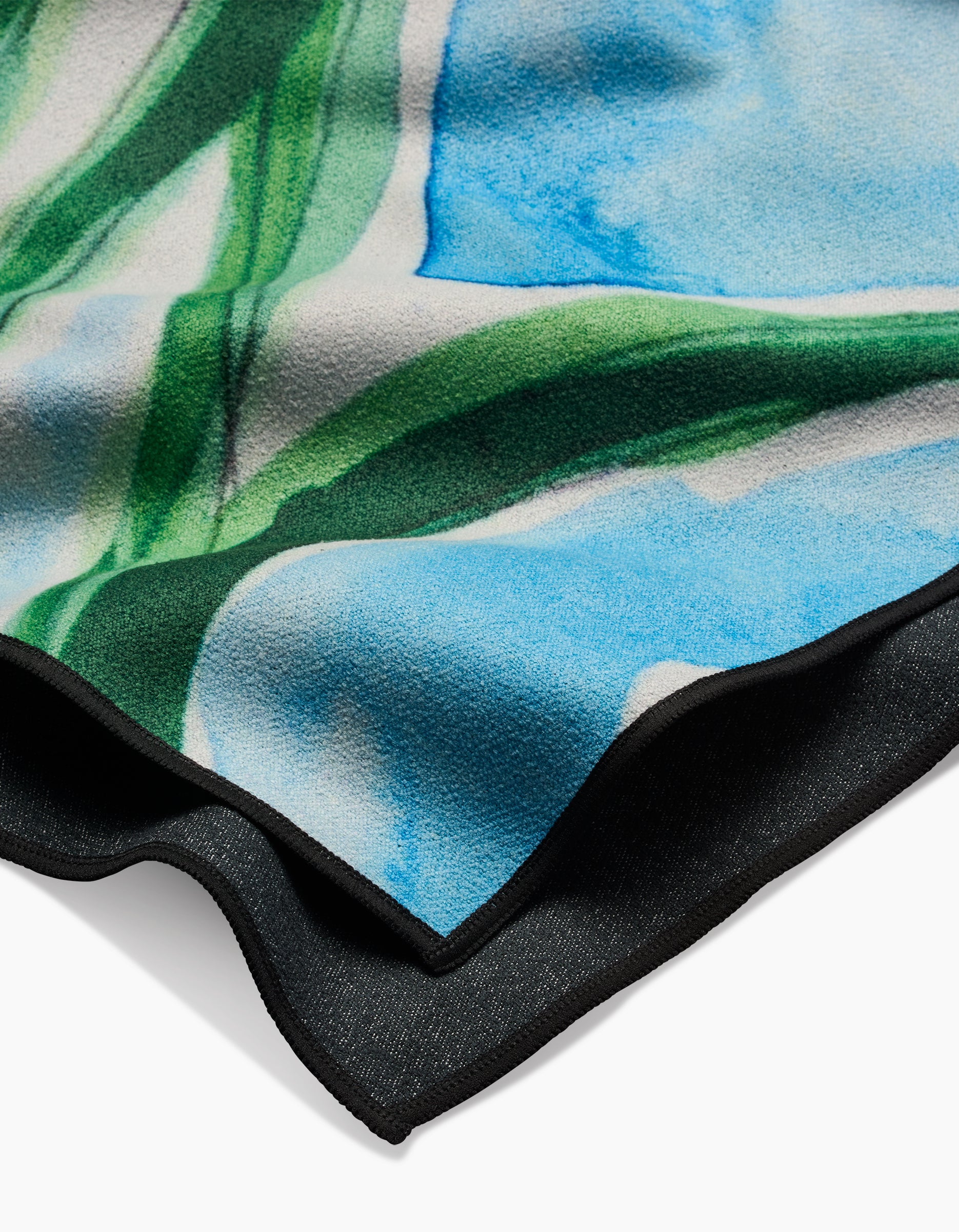 A close-up of the Skye Walker Golden Poppy Flowers beach towel, made from recycled materials, featuring a watercolor-like pattern in blue, green, and white with a black edge, displayed against a white background.