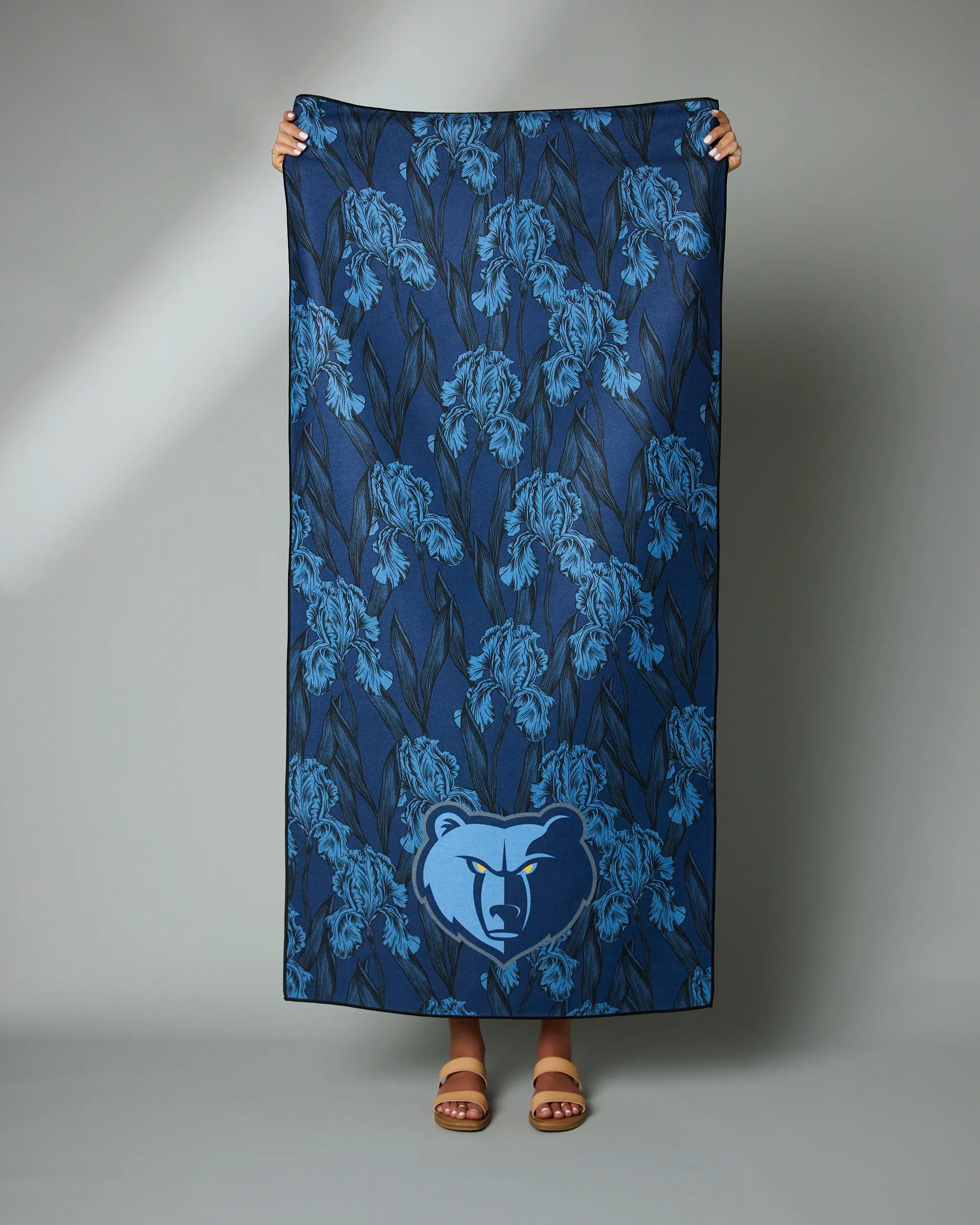 A person displays the Geometry Grizzlies Floral towel, which is blue with a floral print and features a large bear logo at the bottom center. Only their hands, feet, and sandals are visible.