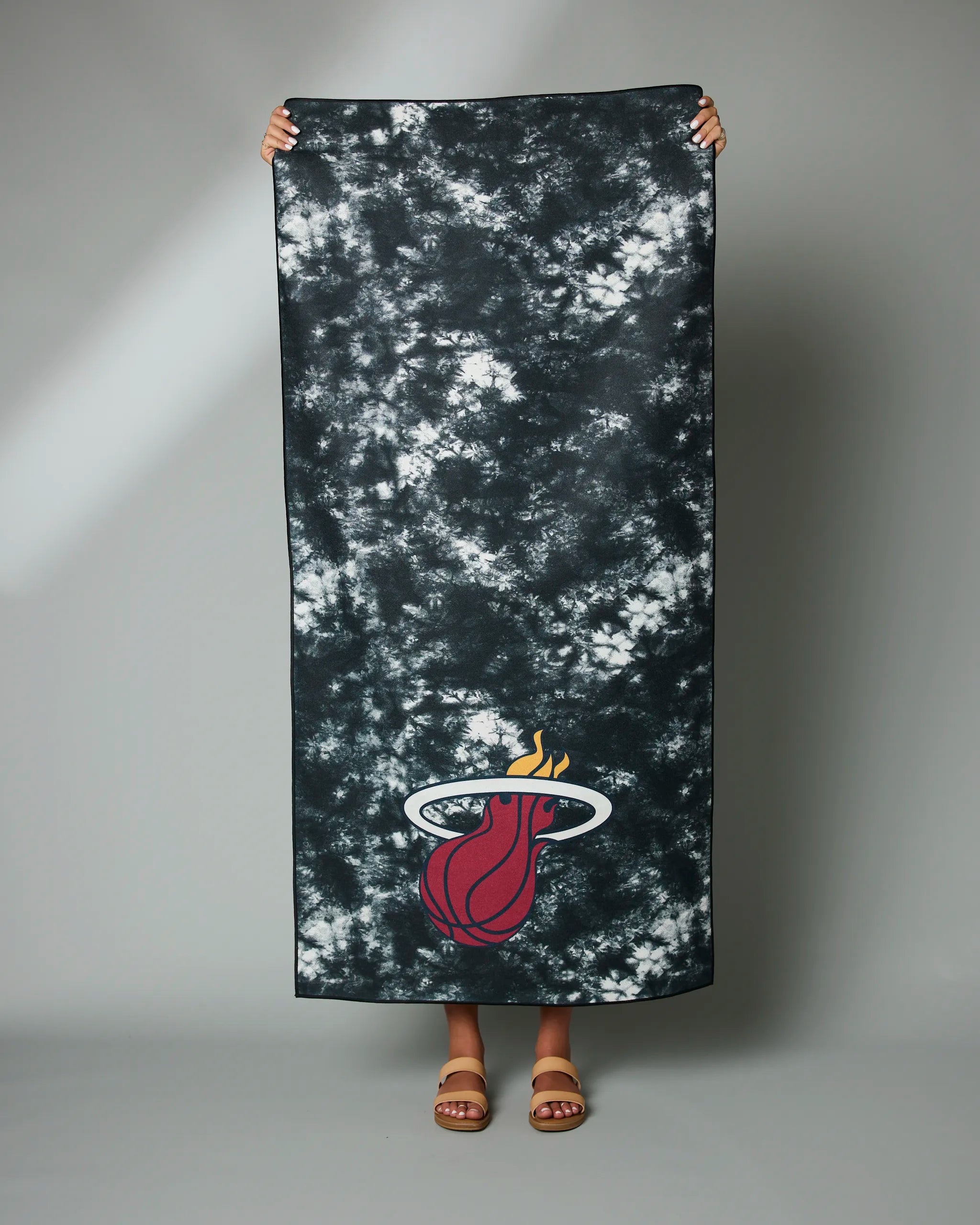 A person displays the Geometry Heat Flash large black and white tie-dye towel featuring the Miami Heat logo, standing against a plain gray background with only their feet in sandals visible.
