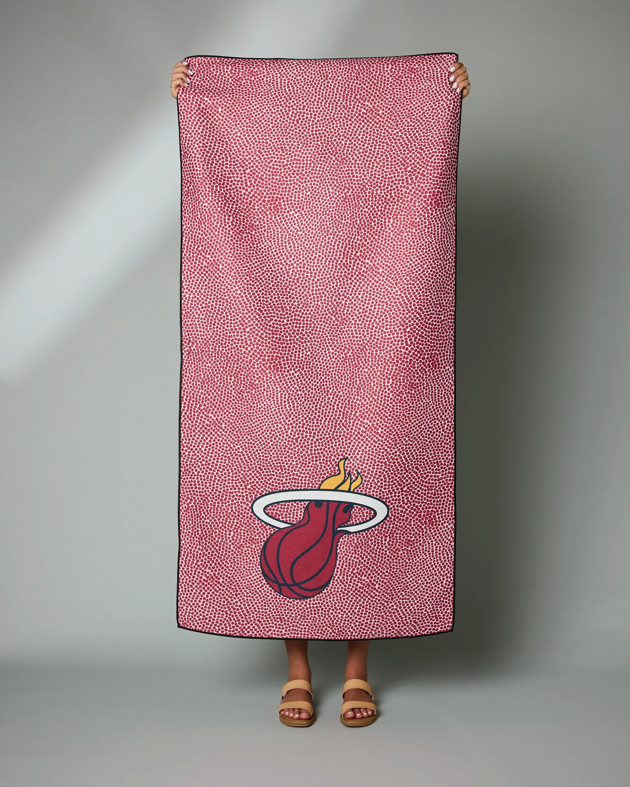 A person holds up the Geometry Heat Grip towel—a rectangular pink towel with a scattered dot pattern and a large Miami Heat logo in the lower right—against a light gray background, showing only their feet and hands.