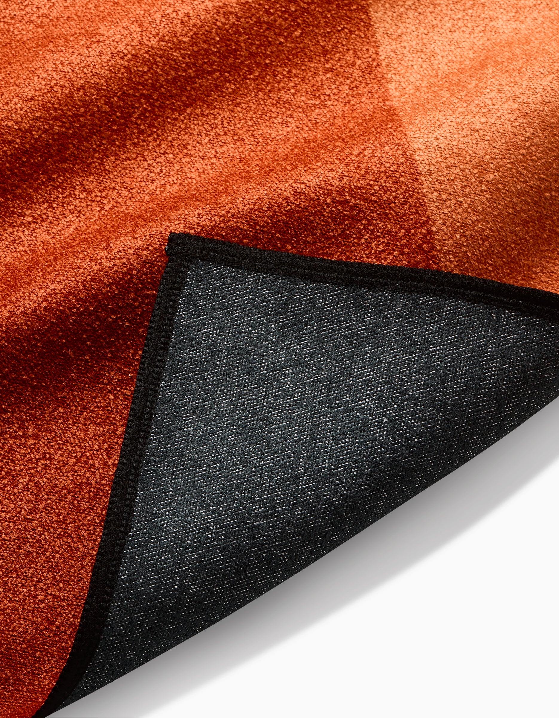 A close-up of Megan Roys In The Desert rug, a reddish-orange velvety piece with black fabric backing and a folded corner revealing its textured underside, crafted from post-consumer recycled materials.