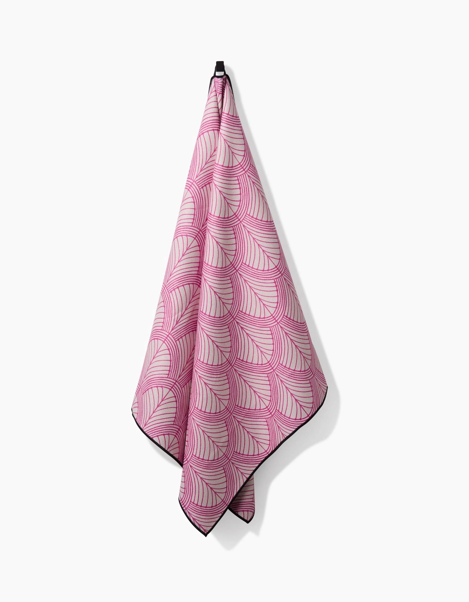 The Amy MacCready Jaskin beach towel features a pink and white curved-line pattern, hangs from a black loop, and is made from quick-drying recycled materials. Shown against a plain white background.