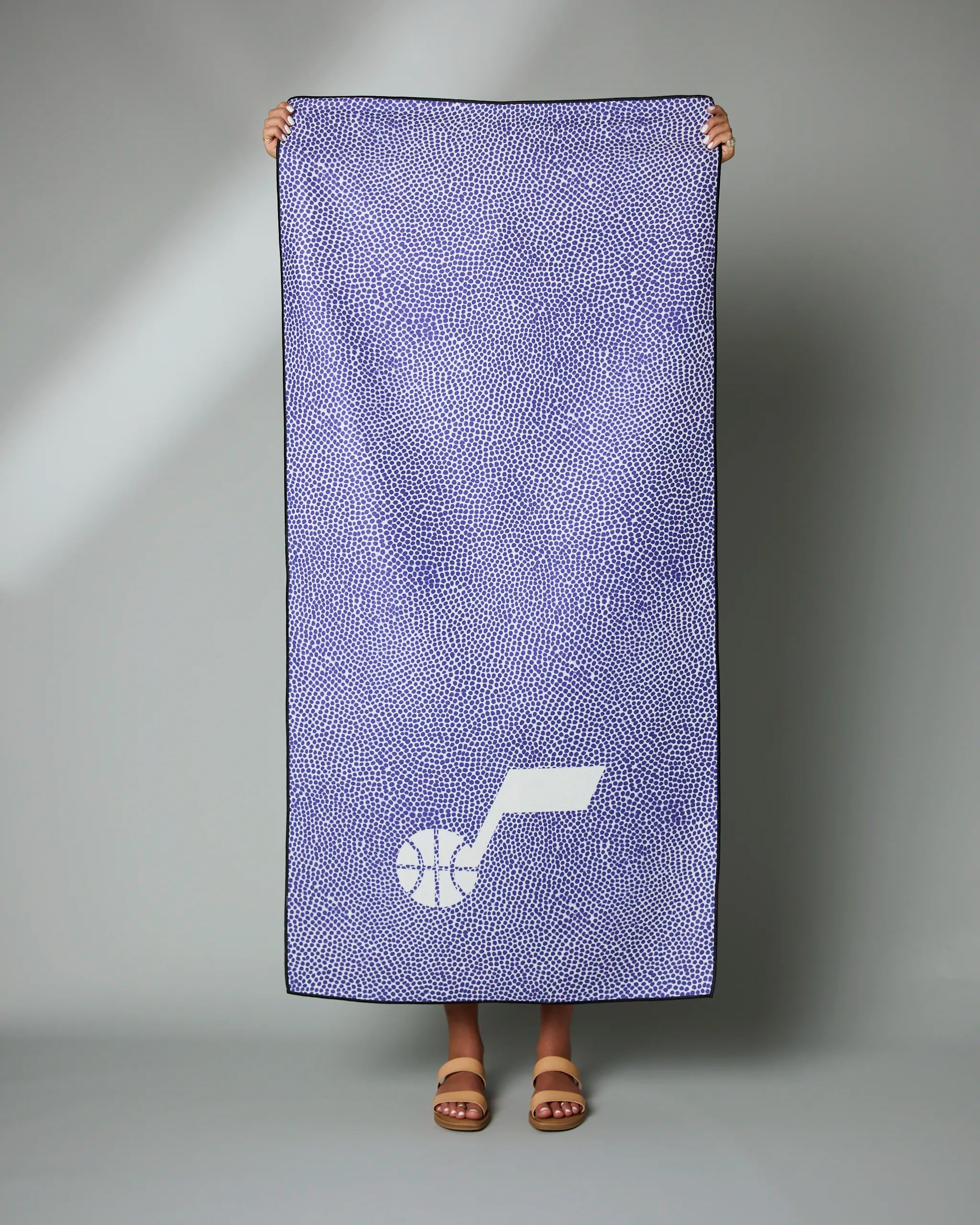 A person stands holding up the Geometry Jazz Grip towel, which is blue and white with a stylized basketball logo in the corner; only their feet in sandals show beneath the towel.