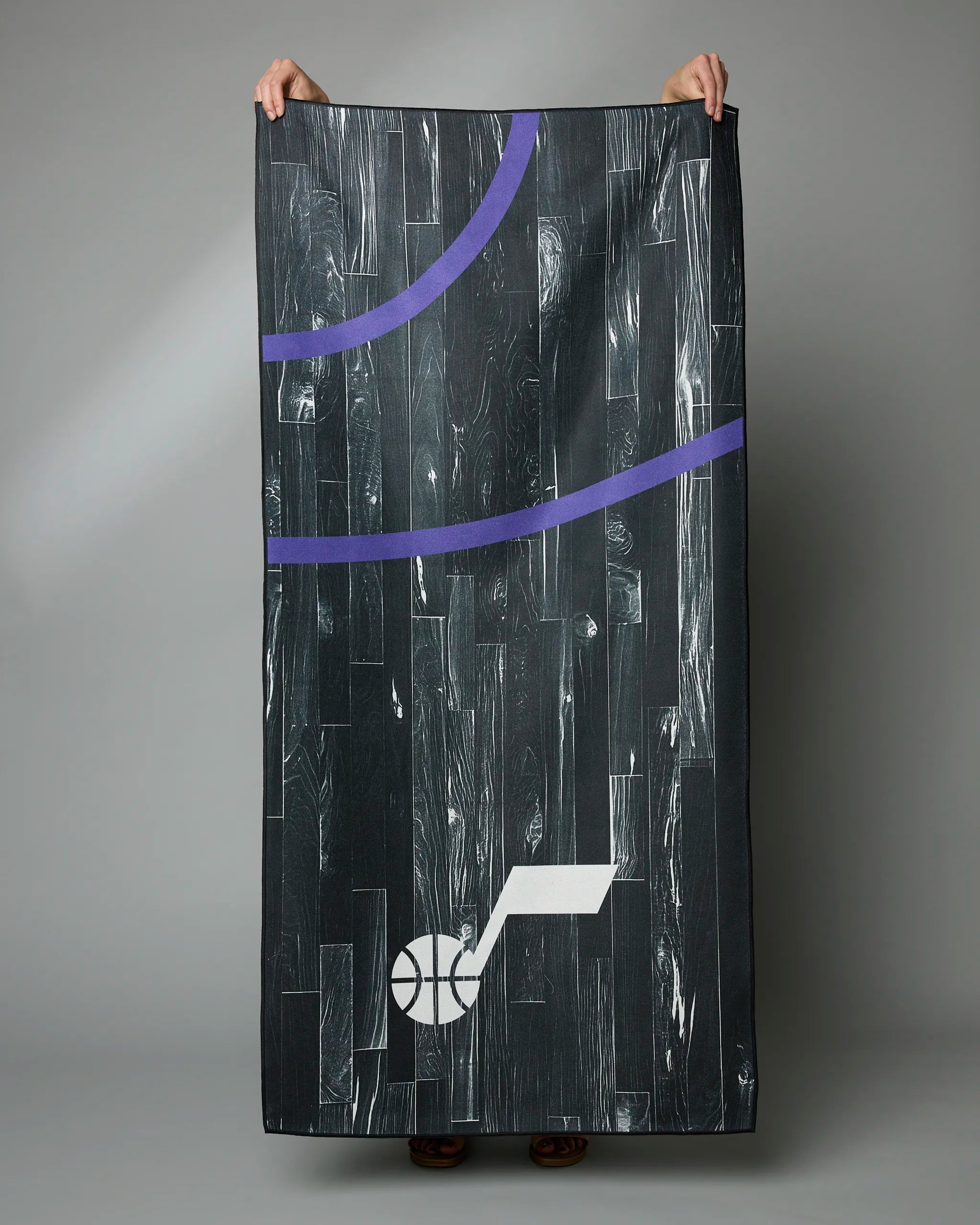 A person holds up the Geometry Jazz Zone Beach Towel, featuring a basketball court design with black wood flooring, curved purple lines, and a white basketball logo—made from recycled materials for a sustainable, quick-drying towel.