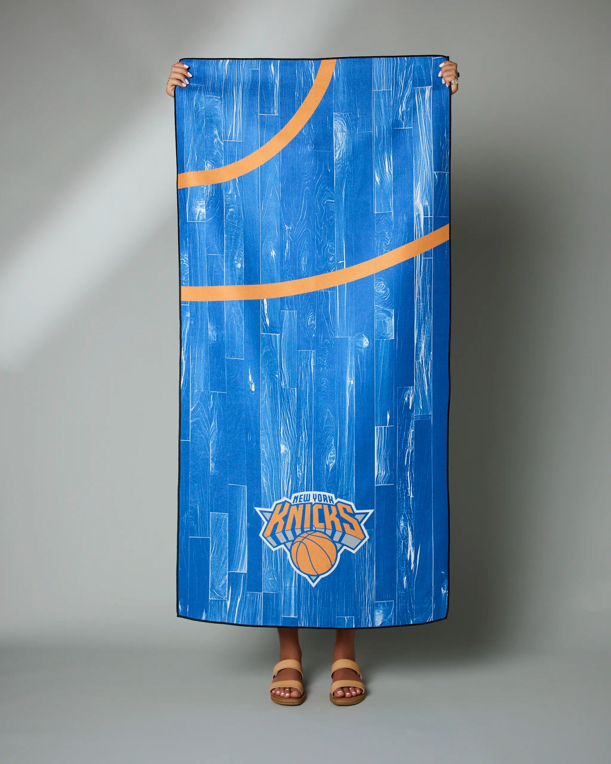 A person holds up the Knicks Zone towel by Geometry, featuring blue fabric with orange basketball court lines and a New York Knicks logo at the bottom; only their hands and sandaled feet are visible.