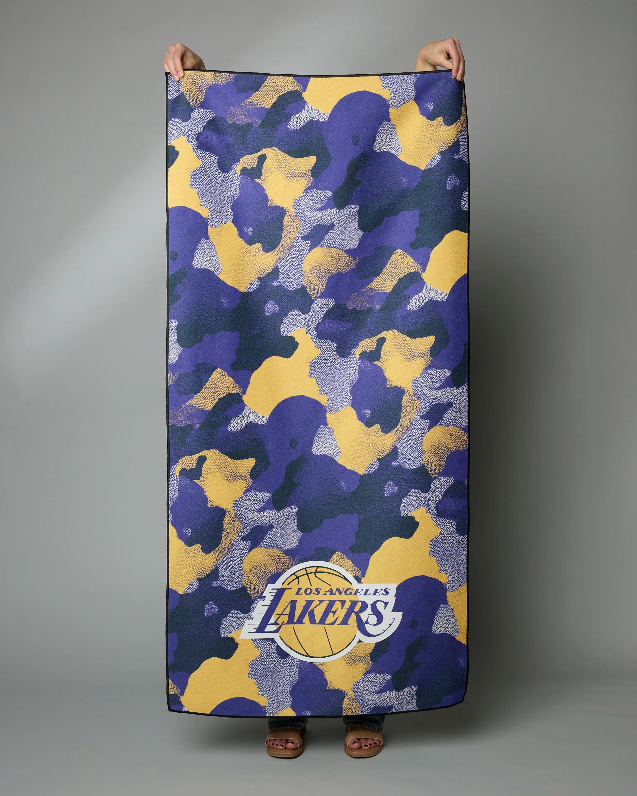 A person holds up the Lakers Camo rectangular micro-loop beach towel by Geometry, featuring a yellow, purple, and blue camouflage pattern with the Los Angeles Lakers logo at the bottom, set against a plain gray background.
