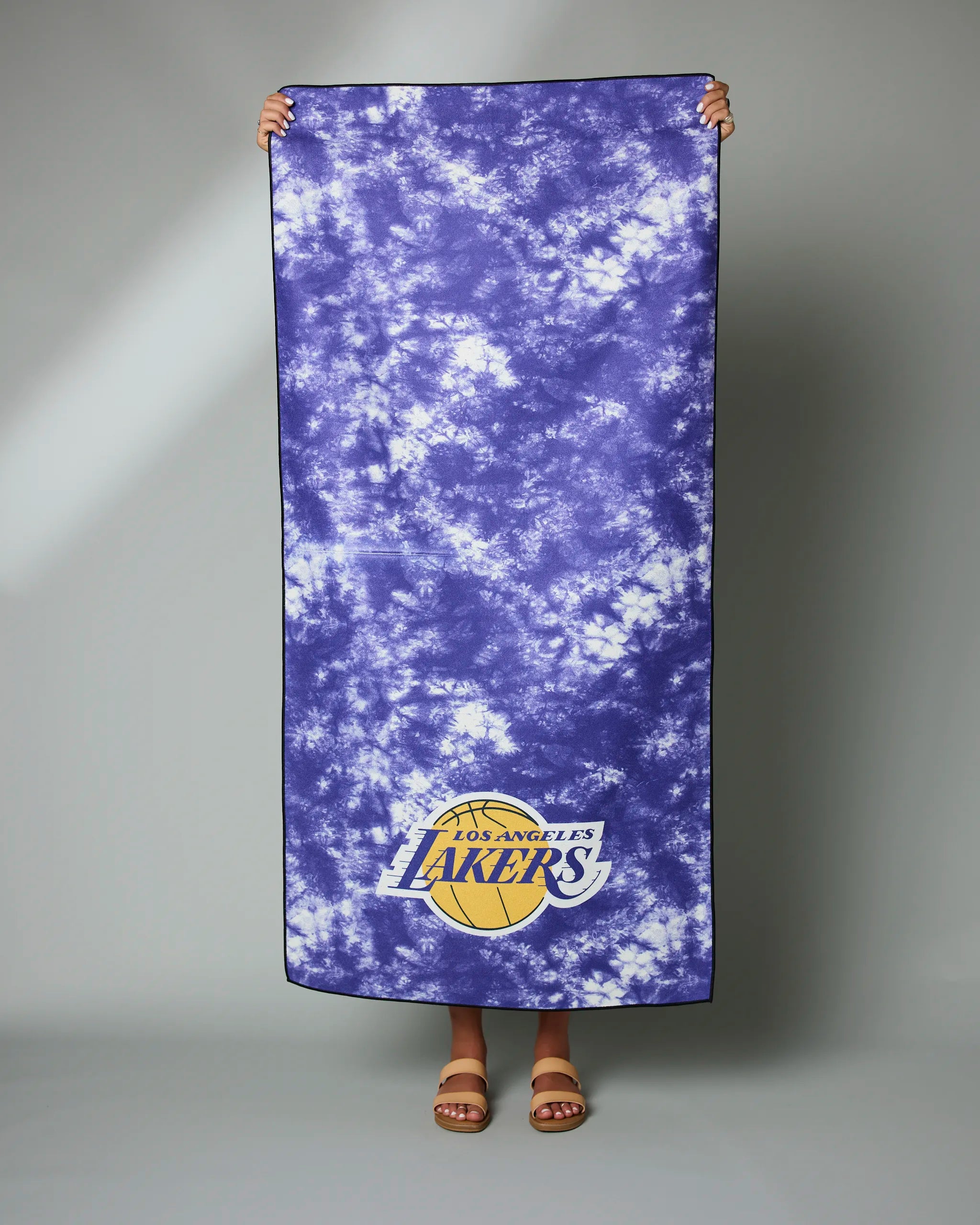 A person holds up the Geometry Lakers Flash towel, a large purple and white tie-dye featuring the Los Angeles Lakers logo at the bottom. Only their hands and feet are visible.