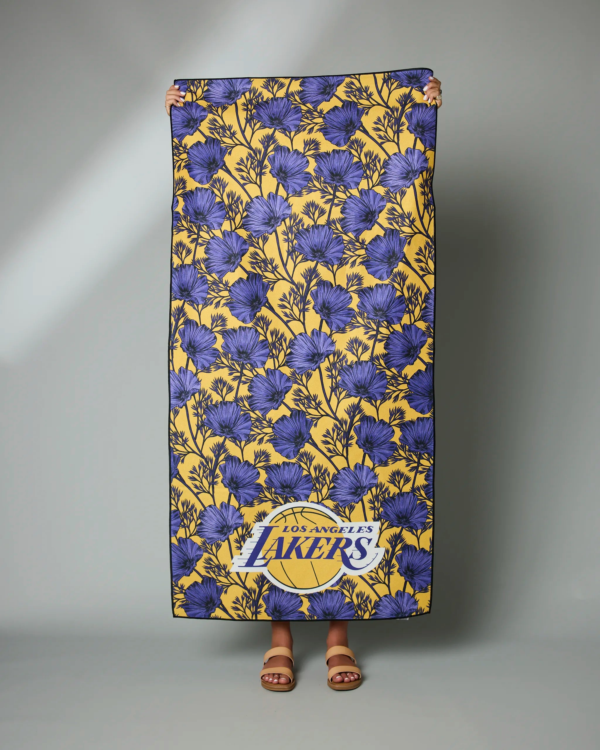A person holds up the Geometry Lakers Floral beach towel, featuring a large yellow and blue floral design with the Los Angeles Lakers logo. Only the hands and feet in brown sandals are visible.