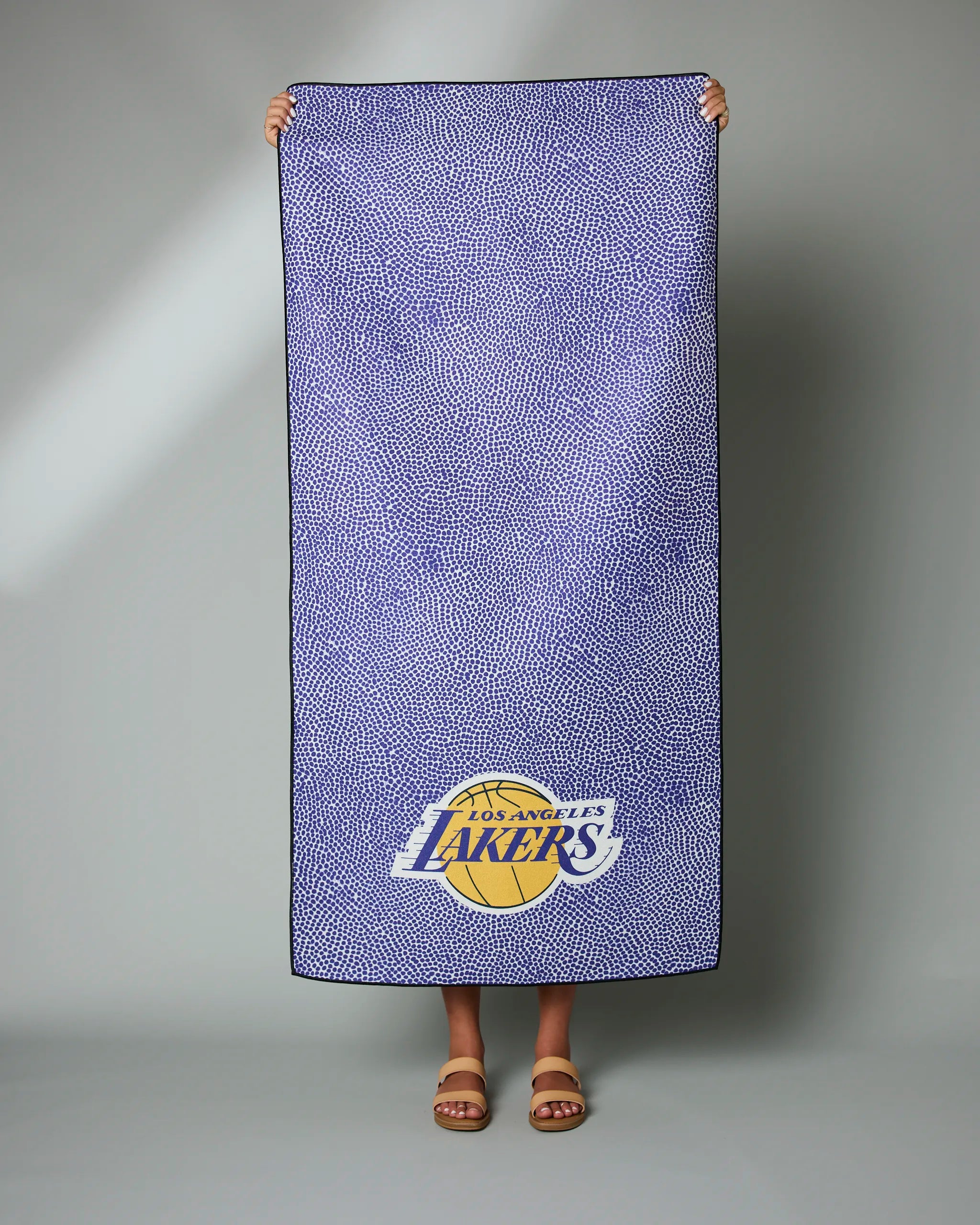 Someone holds up the Geometry Lakers Grip, a rectangular blue towel with small white dots and the Los Angeles Lakers logo in the bottom right corner. Only their hands and feet are visible.