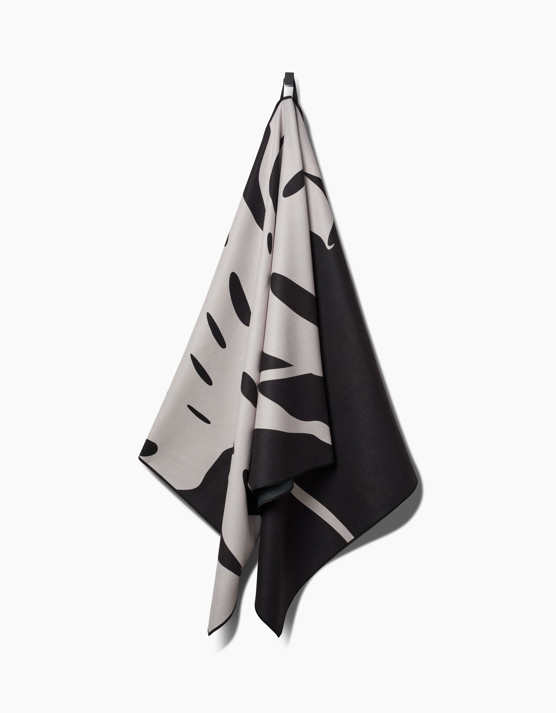 A quick-drying PrintedPlant Monstera Micro-loop Beach Towel with a bold, abstract black and beige pattern hangs on a hook against a plain white background.