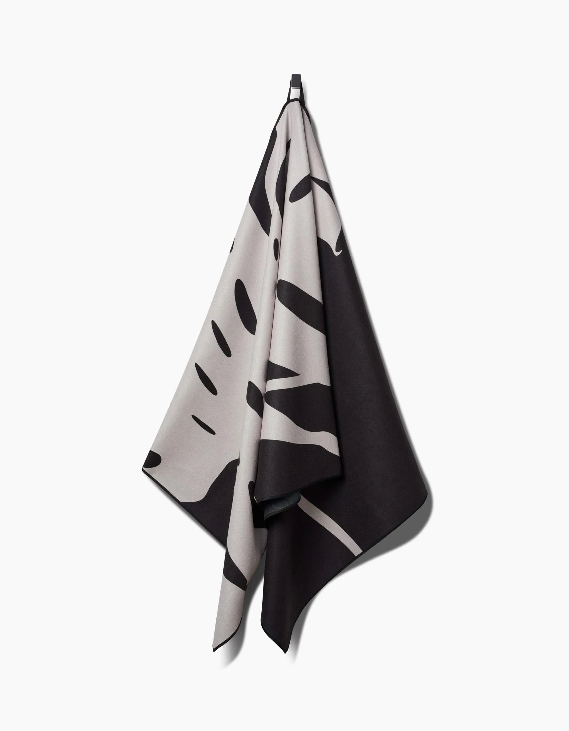 A quick-drying PrintedPlant Monstera Micro-loop Beach Towel with a bold, abstract black and beige pattern hangs on a hook against a plain white background.