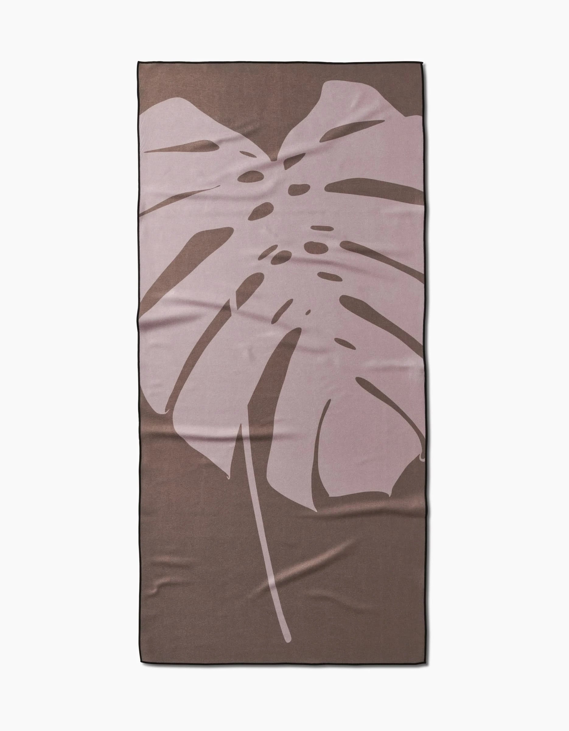 The Monstera Leaf beach towel by PrintedPlant is rectangular and brown with a large light pink monstera leaf in the center. Made from quick-drying recycled materials, it has neatly finished edges and a soft, smooth surface.